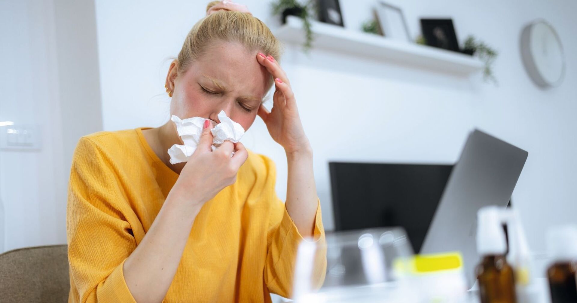 Eine Frau mit blonden Haaren niest in ein Taschentuch. Ihr Gesicht ist gerötet. Sie trägt einen gelber Pullover. Am Tisch vor ihr ist ein Laptop und verschiedene Medikamente zu sehen. An der Wand ist ein Uhr und ein Regal auf dem Bilder stehen montiert.