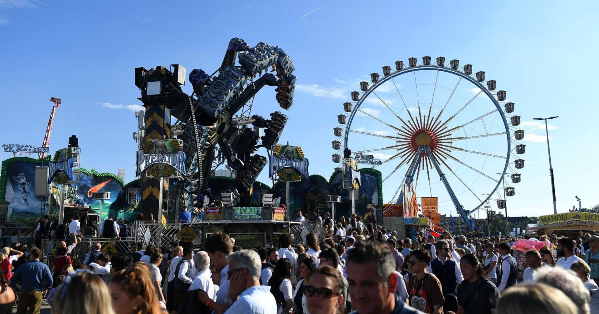 Menschenmenge auf dem Oktoberfest in München, im Hintergrund ein großes Riesenrad und Fahrgeschäfte bei blauem Himmel. Leiche hinter dem Riesenrad auf dem Oktoberfest in München entdeckt – Polizei vermutet medizinischen Notfall.