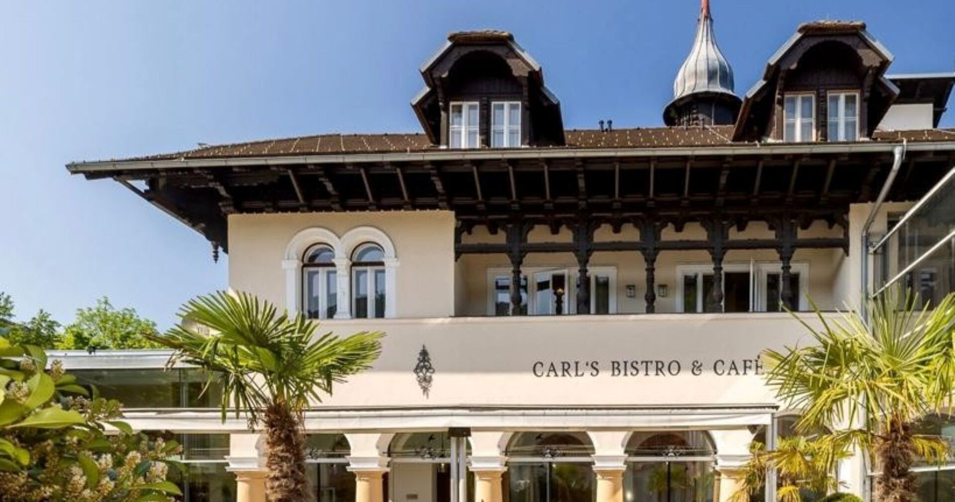 Fassade des Hotel Sacher in Baden mit dem Schriftzug „Carl’s Bistro &amp; Café“, flankiert von Palmen, unter blauem Himmel.