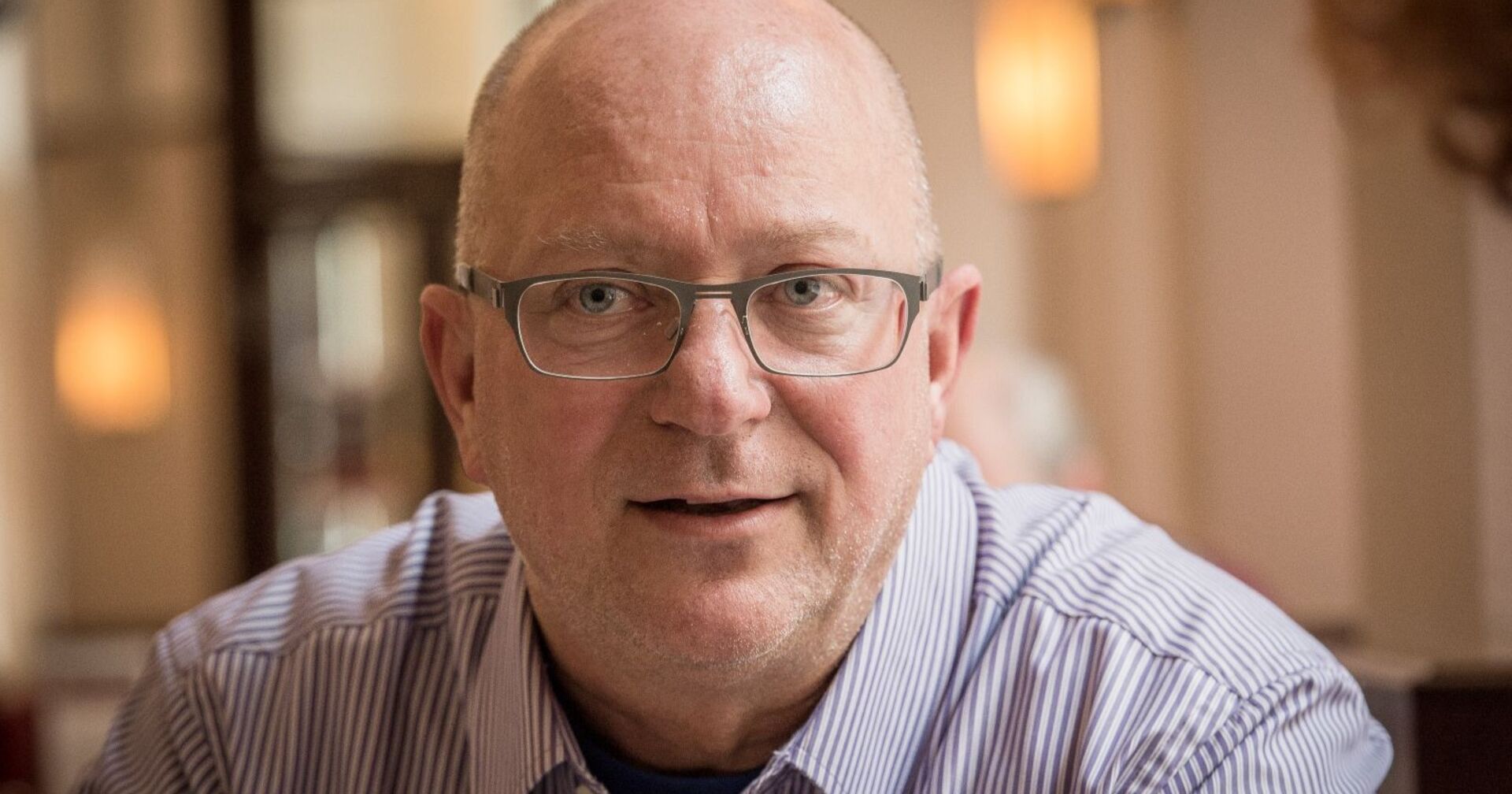 Porträt von Niki Glattauer mit Brille und gestreiftem Hemd in einem Café, frontal aufgenommen, leichtes Lächeln, verschwommener Hintergrund mit warmem Licht.