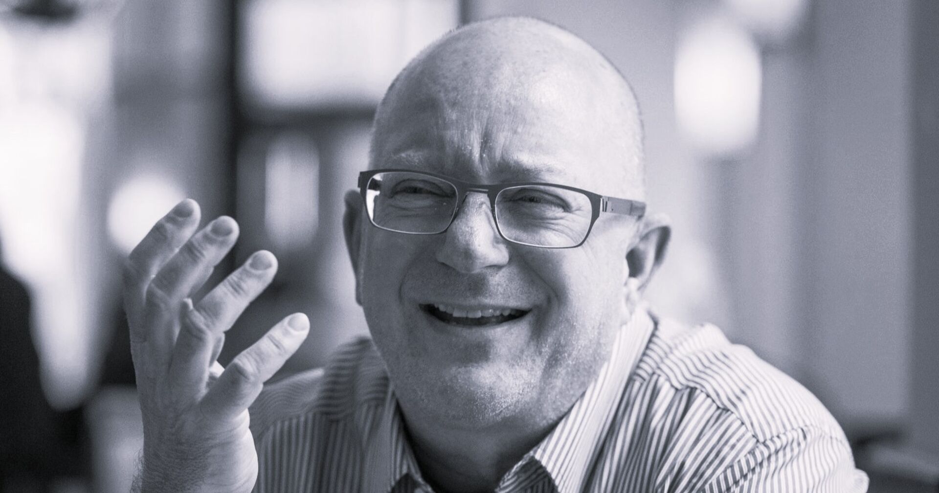 Schwarz-weiß-Porträt von Niki Glattauer, lachend, mit Brille und gestreiftem Hemd, hebt eine Hand gestikulierend.