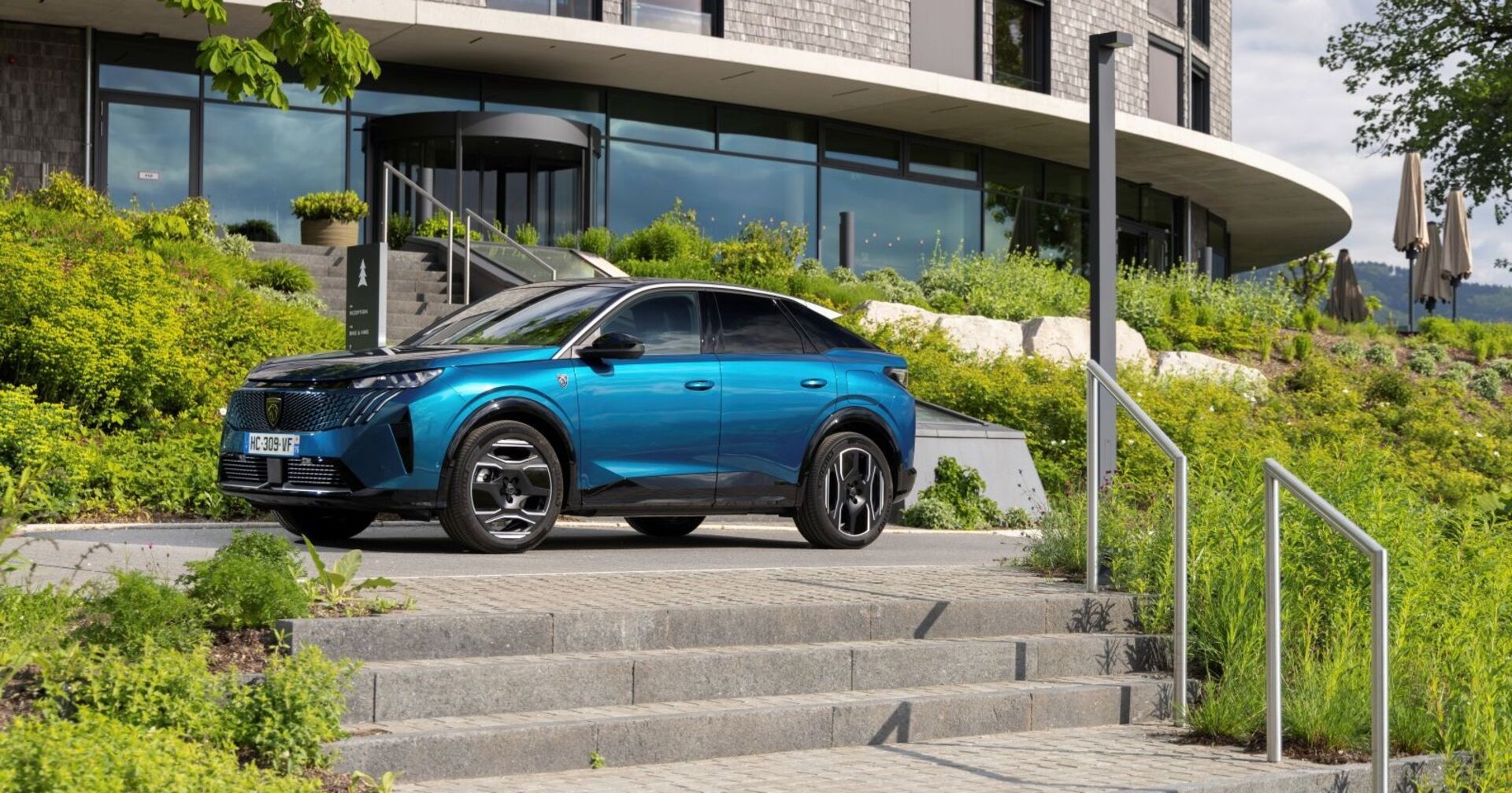 Ein blauer Peugeot E-3008 parkt am vor einer Treppe. Im Hintergrund ist ein Haus zu sehen. Links und rechts vom Auto wachsen einige Sträucher.