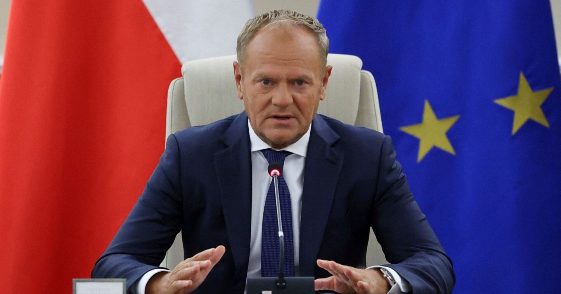 Donald Tusk sitzt in einem dunklen Anzug vor Mikrofonen zwischen einer polnischen und einer EU-Flagge bei einer Krisensitzung.