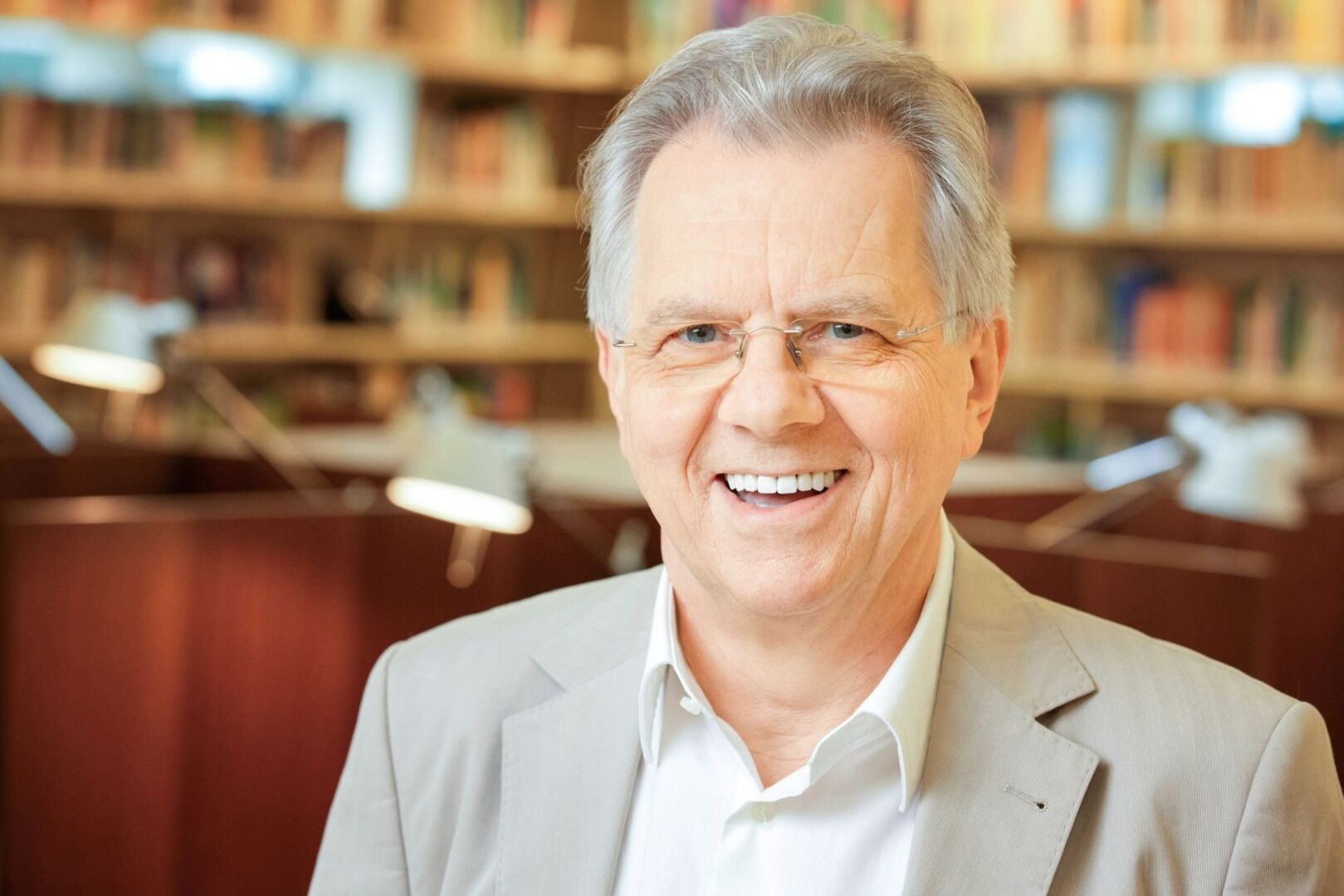 Zukunftsforscher Reinhold Popp in weißem Hemd und beigem Sakko, mit Brille lächelt in die Kamera. Im Hintergrund sieht man verschwommen Regale mit jede Menge Büchern.