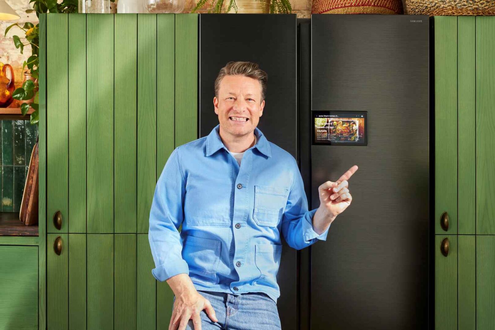 Koch Jamie Oliver steht lächelnd in blauem Hemd vor einer grünen Küche mit modernem Kühlschrank, in dem ein Tablet integriert ist, das Rezepte anzeigt.