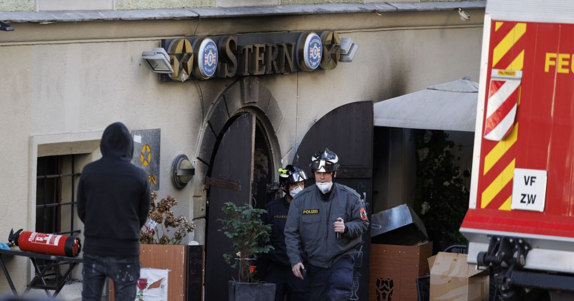 Polizisten mit Atemschutzmasken verlassen das ausgebrannte Lokal „Stern“ in der Grazer Sporgasse, davor beschädigte Einrichtung und ein Feuerlöscher.