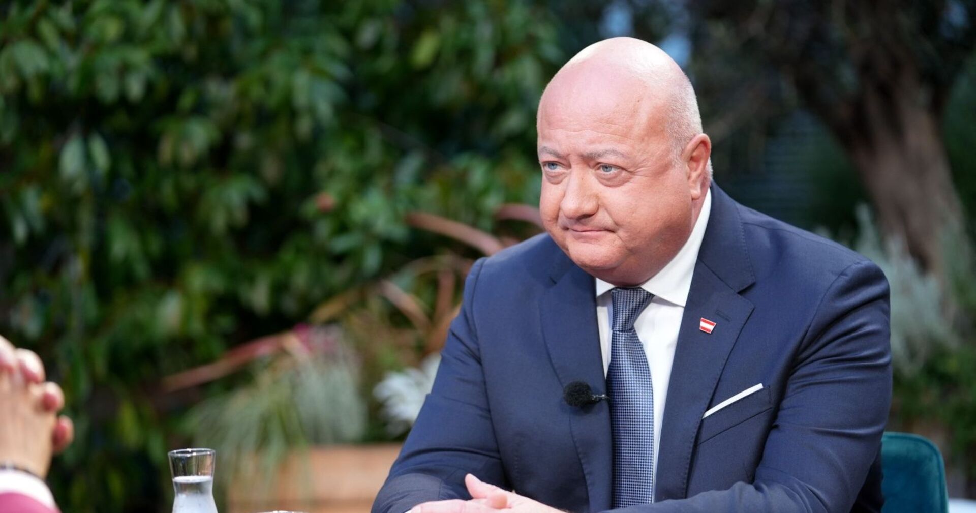 Christian Stocker im dunklen Anzug mit Krawatte und Österreich-Anstecker sitzt bei einem Fernsehinterview im Freien. Vor ihm steht ein Glas Wasser, im Hintergrund sind Pflanzen zu sehen.