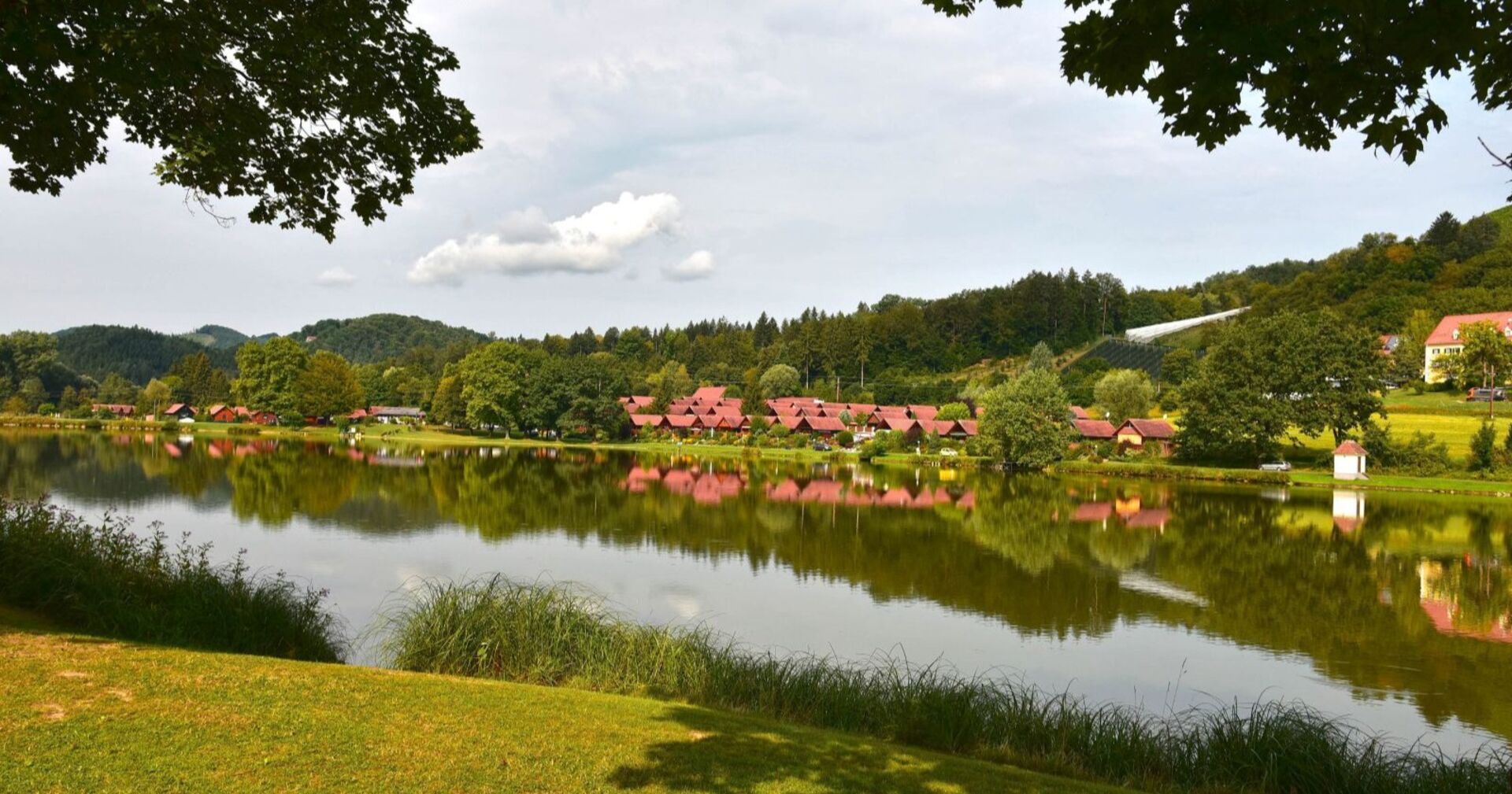 Blick über den Sulmsee in der Steiermark, mit spiegelglattem Wasser, roten Hütten am Ufer und dicht bewaldeten Hügeln im Hintergrund.