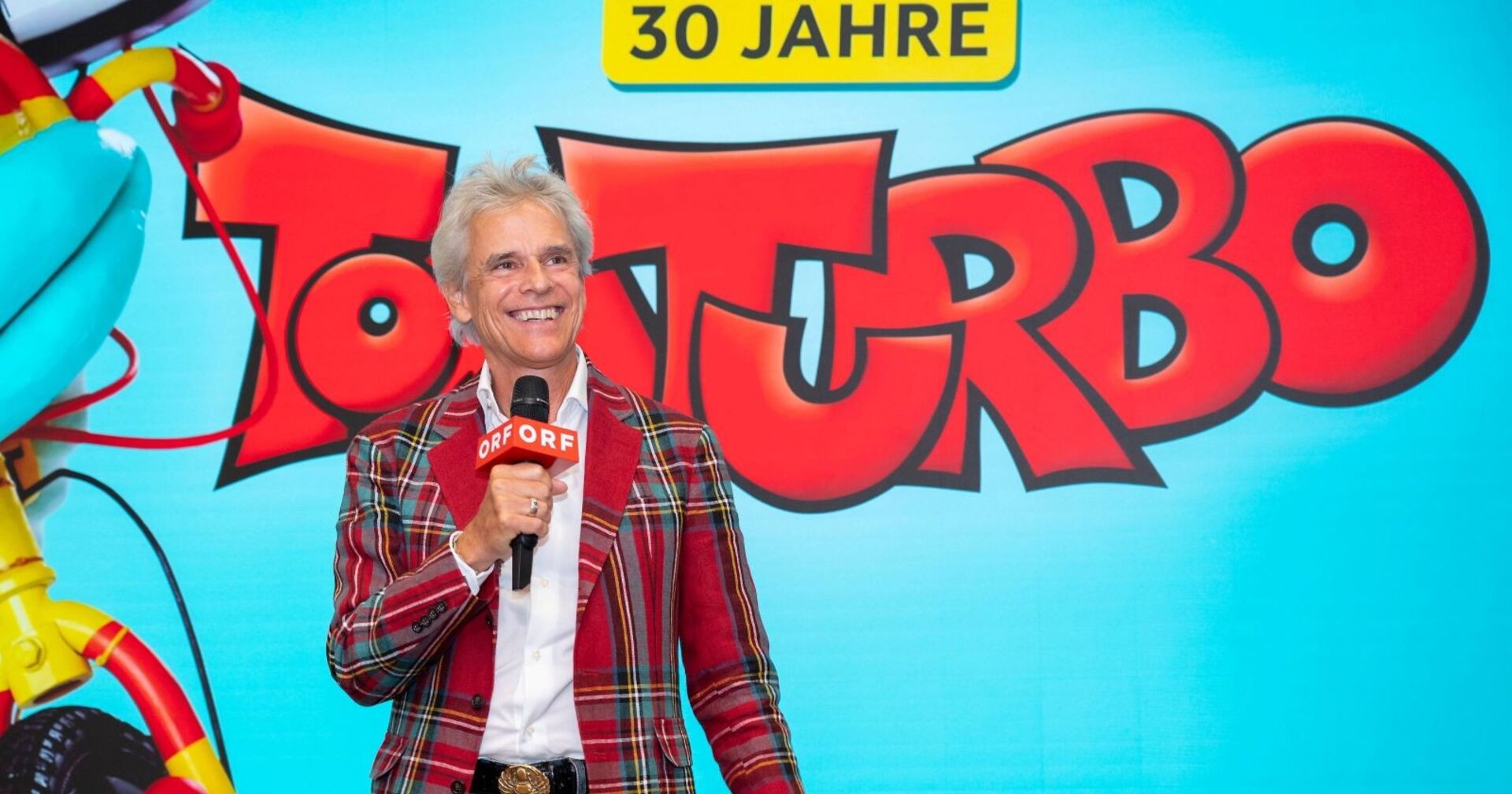 Thomas Brezina hält ein ORF-Mikrofon, lächelt vor einer großen „Tom Turbo“-Jubiläumswand mit der Aufschrift „30 Jahre“.