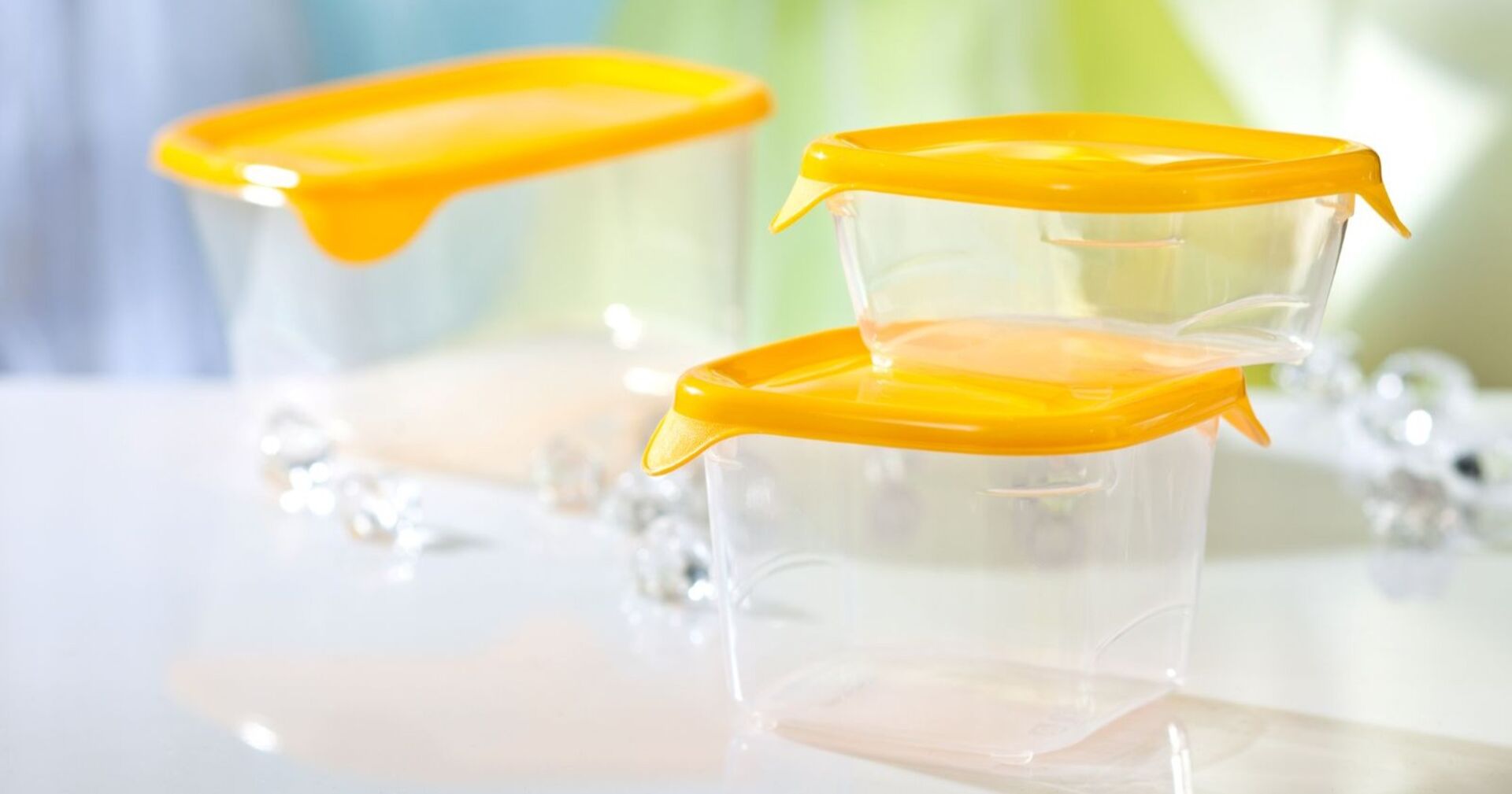 Drei durchsichtige Frischhaltedosen aus Kunststoff mit gelben Deckeln, übereinander gestapelt auf einem hellen Untergrund, im Hintergrund verschwommene Farben in Grün und Blau.