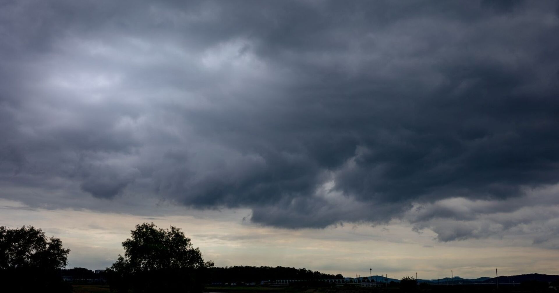 Dunkle, tiefhängende Gewitterwolken ziehen über eine Landschaft mit Bäumen und Hügeln im Hintergrund – Vorbote eines schweren Unwetters.