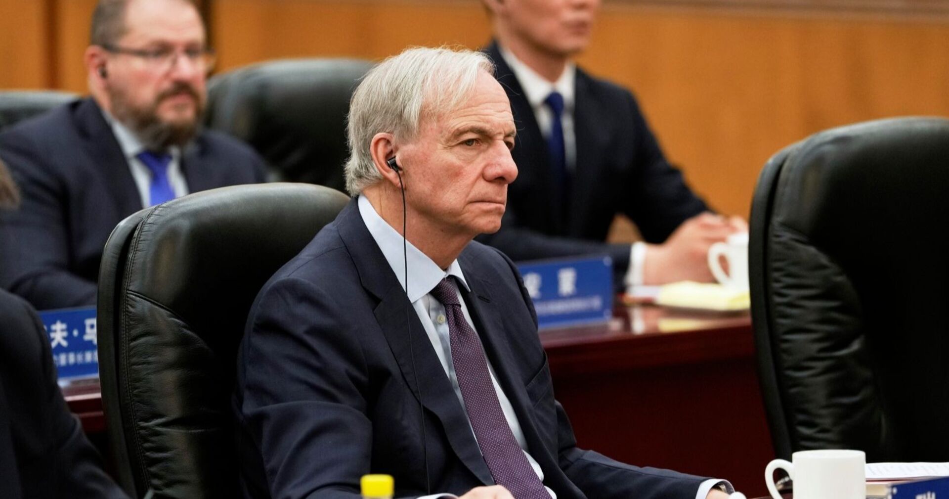 Ray Dalio sitzt im Anzug mit Kopfhörern bei einer Konferenz am Tisch, konzentrierter Gesichtsausdruck, weitere Teilnehmer im Hintergrund.