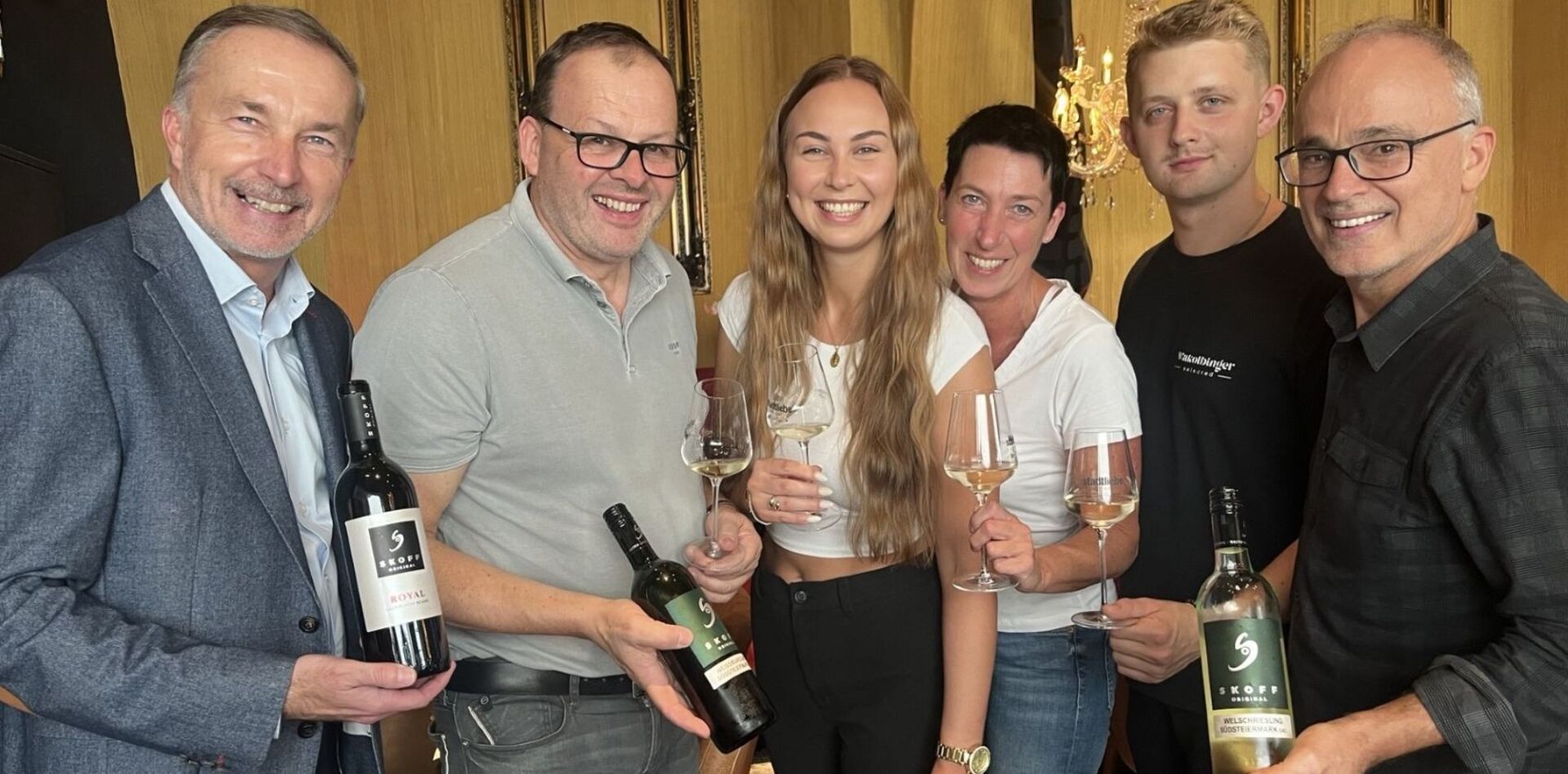 Vier Männer und zwei Männer lachen in die Kamera. In den Händen halten sie Weinflaschen beziehungsweise Weingläser, die mit Weißwein gefüllt sind.