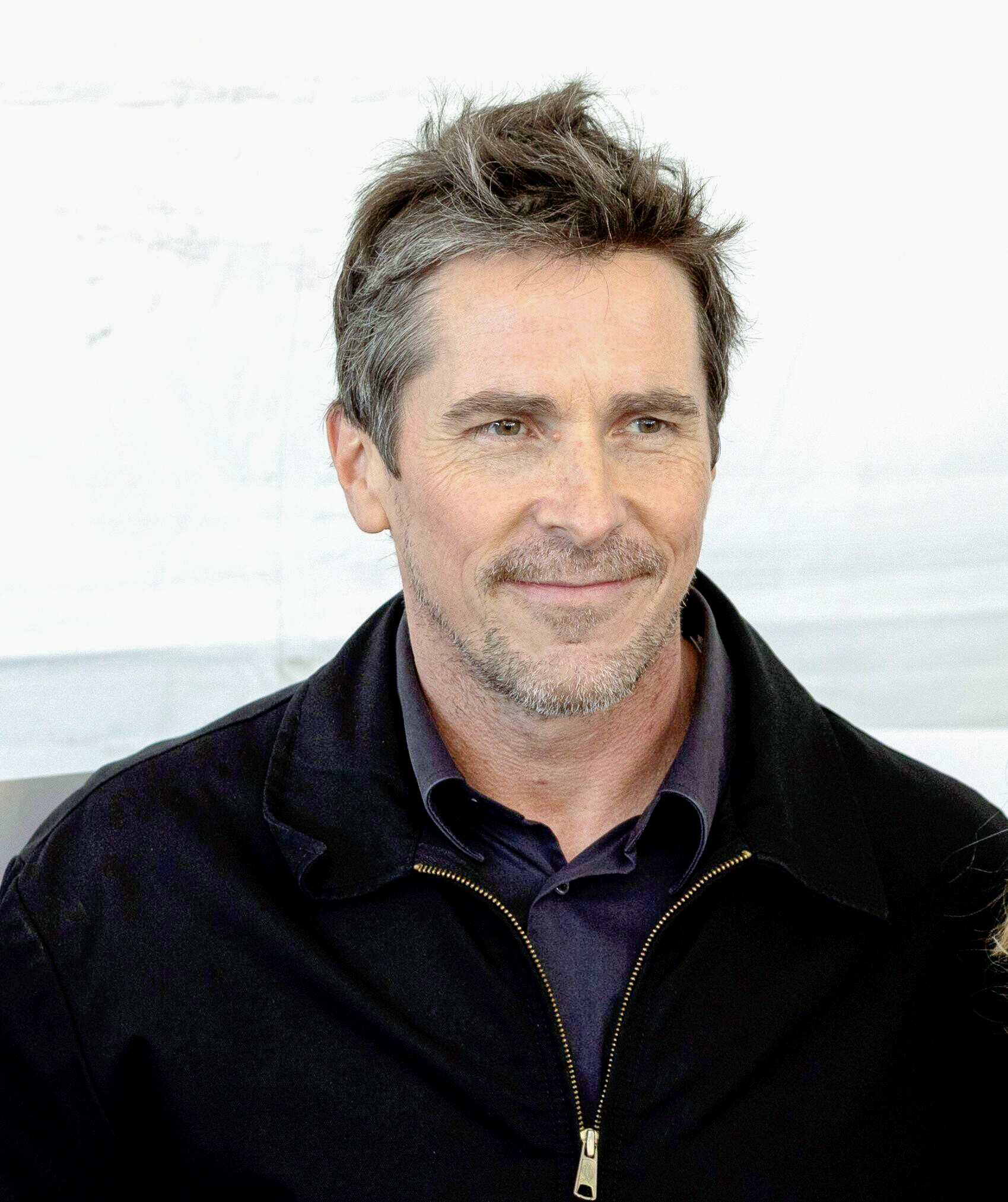 Christian Bale in dunkler Jacke und Hemd, lächelnd.