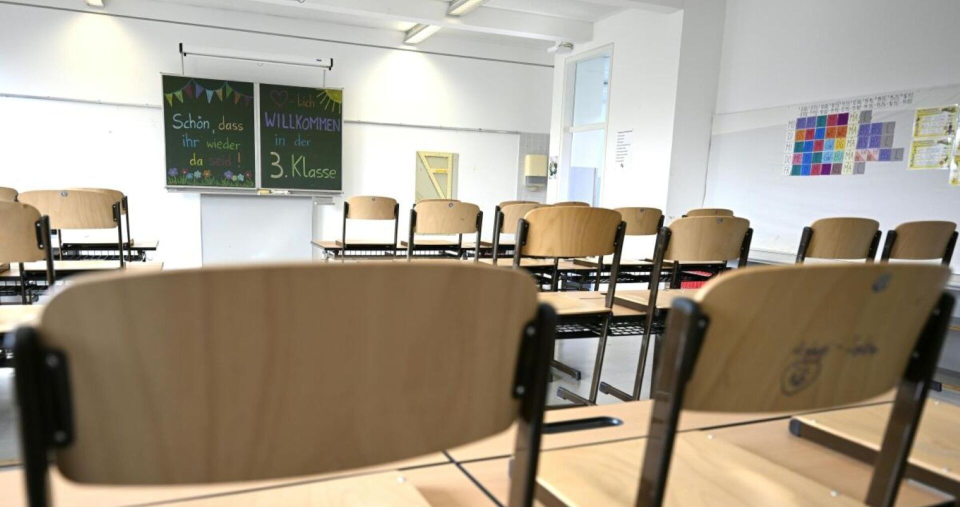 Leeres Klassenzimmer mit aufgestellten Sesseln und Tafelaufschrift „Willkommen in der 3. Klasse“.