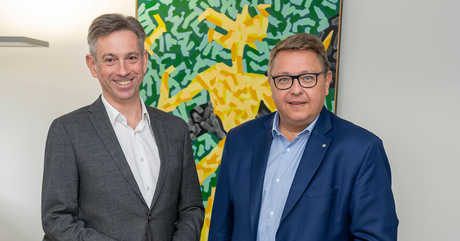 Werner Ressi und Martin Graf stehen nebeneinander vor einem farbenfrohen abstrakten Gemälde und lächeln in die Kamera.