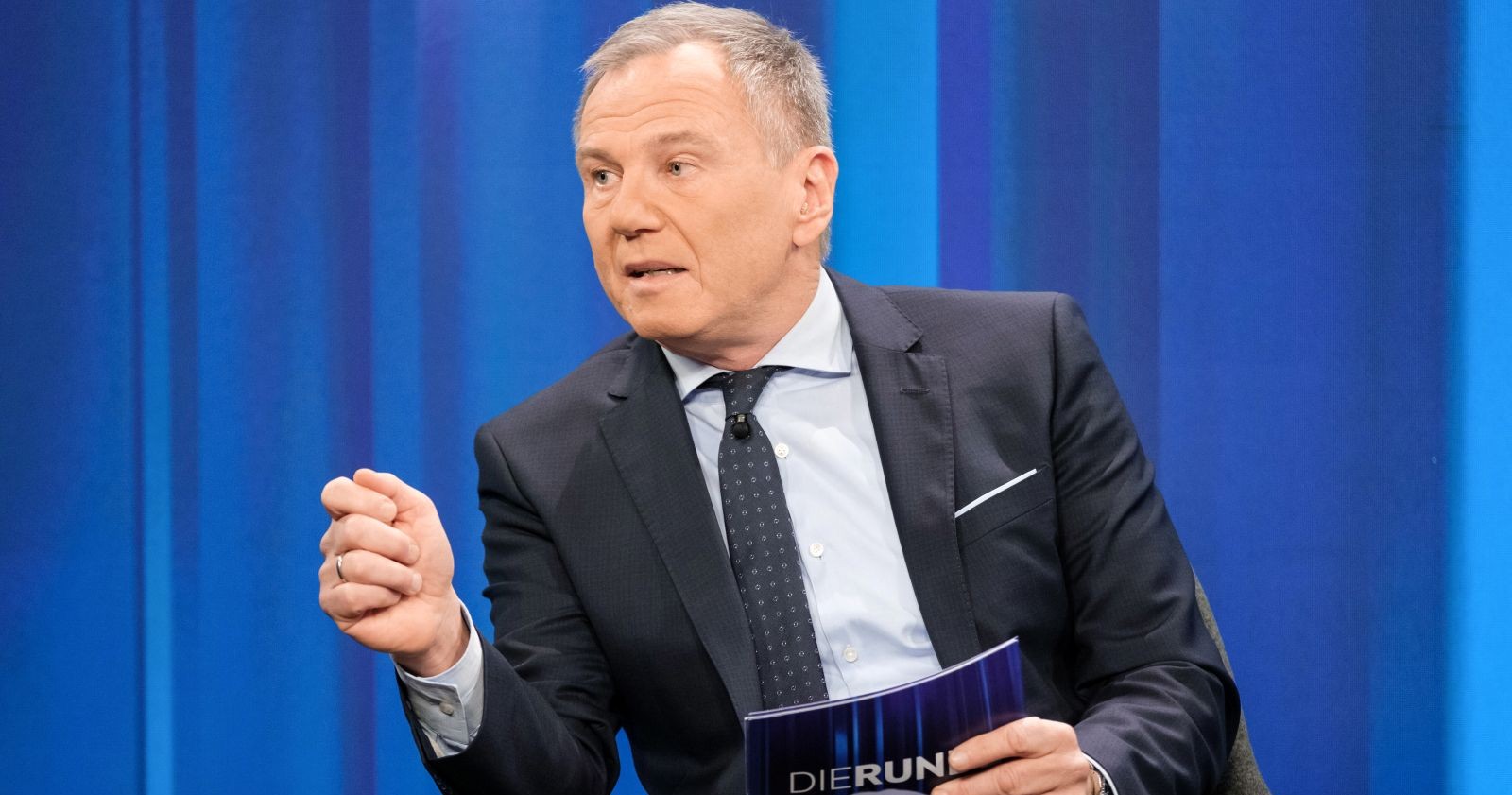 Armin Wolf sitzt in einem TV-Studio vor blauem Hintergrund und gestikuliert während eines Gesprächs.