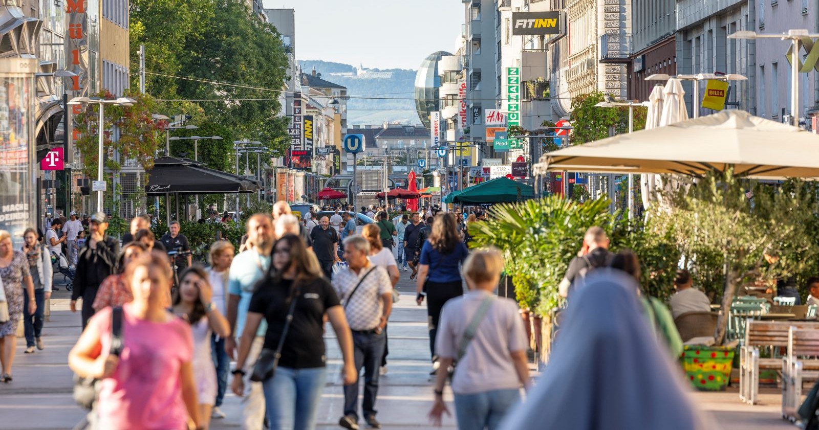 Menschen spazieren und shoppen auf einer belebten Einkaufsstraße in Wien mit Geschäften, Cafés und U-Bahn-Schildern im Hintergrund.