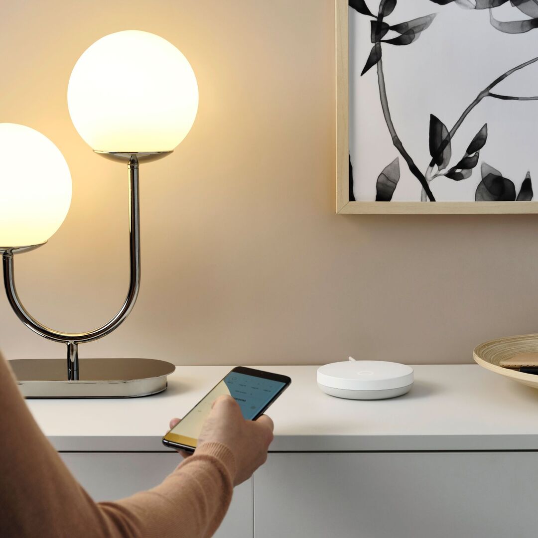 Person steuert per Smartphone einen modernen Bewegungssensor im Smart Home; daneben eine Designlampe und stilvolle Wohnzimmereinrichtung.
