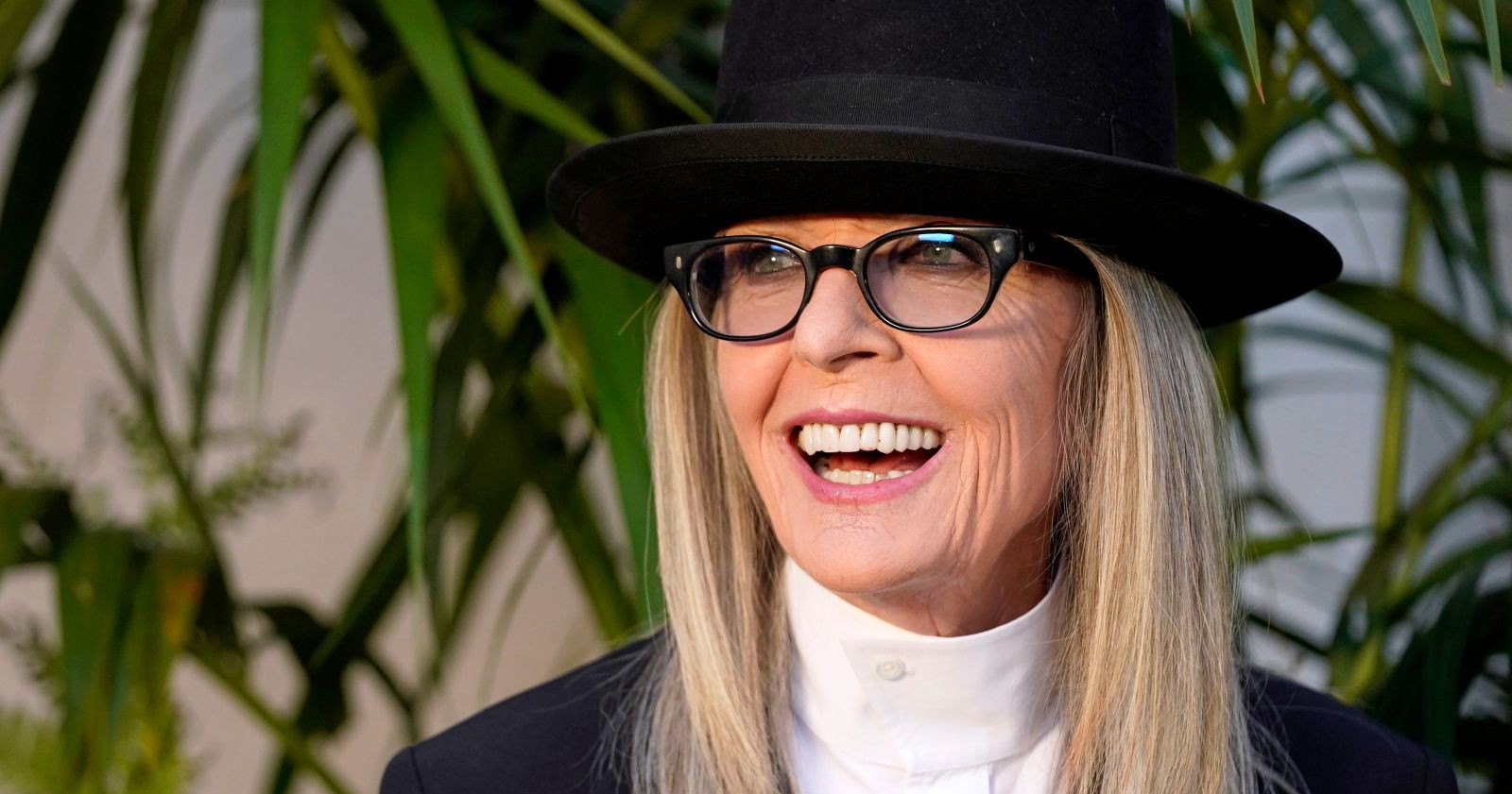 Diane Keaton, lachend, trägt einen schwarzen Hut und eine Brille; sie steht vor grünen Pflanzen.