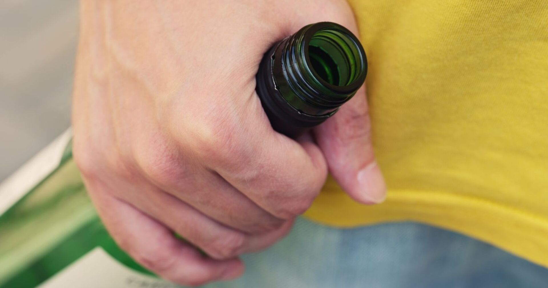 Mann mit gelbem T-Shit hält in aggressiver Grundhaltung Weinflasche aus Grünglas in seiner rechten Hand