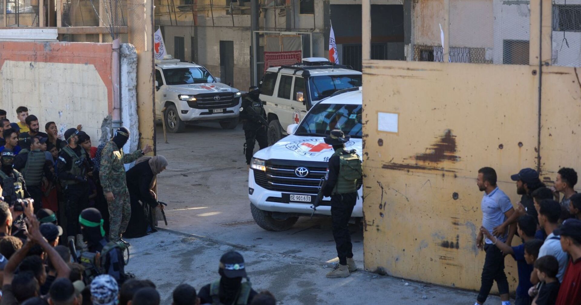 Weiße Fahrzeuge des Roten Kreuzes fahren durch ein Tor im Gazastreifen, bewaffnete Kämpfer sichern die Szene, während Menschen am Straßenrand zusehen.