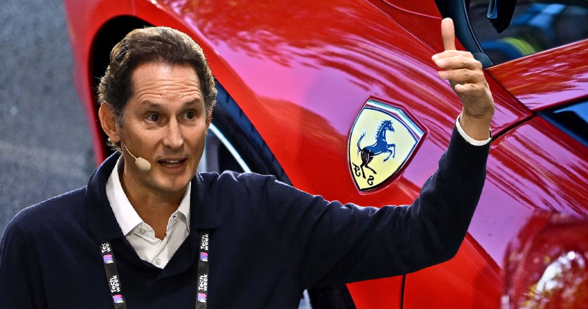 John Elkann bei einer Präsentation, im Hintergrund der Ausschnitt eines knallroten Ferraris.