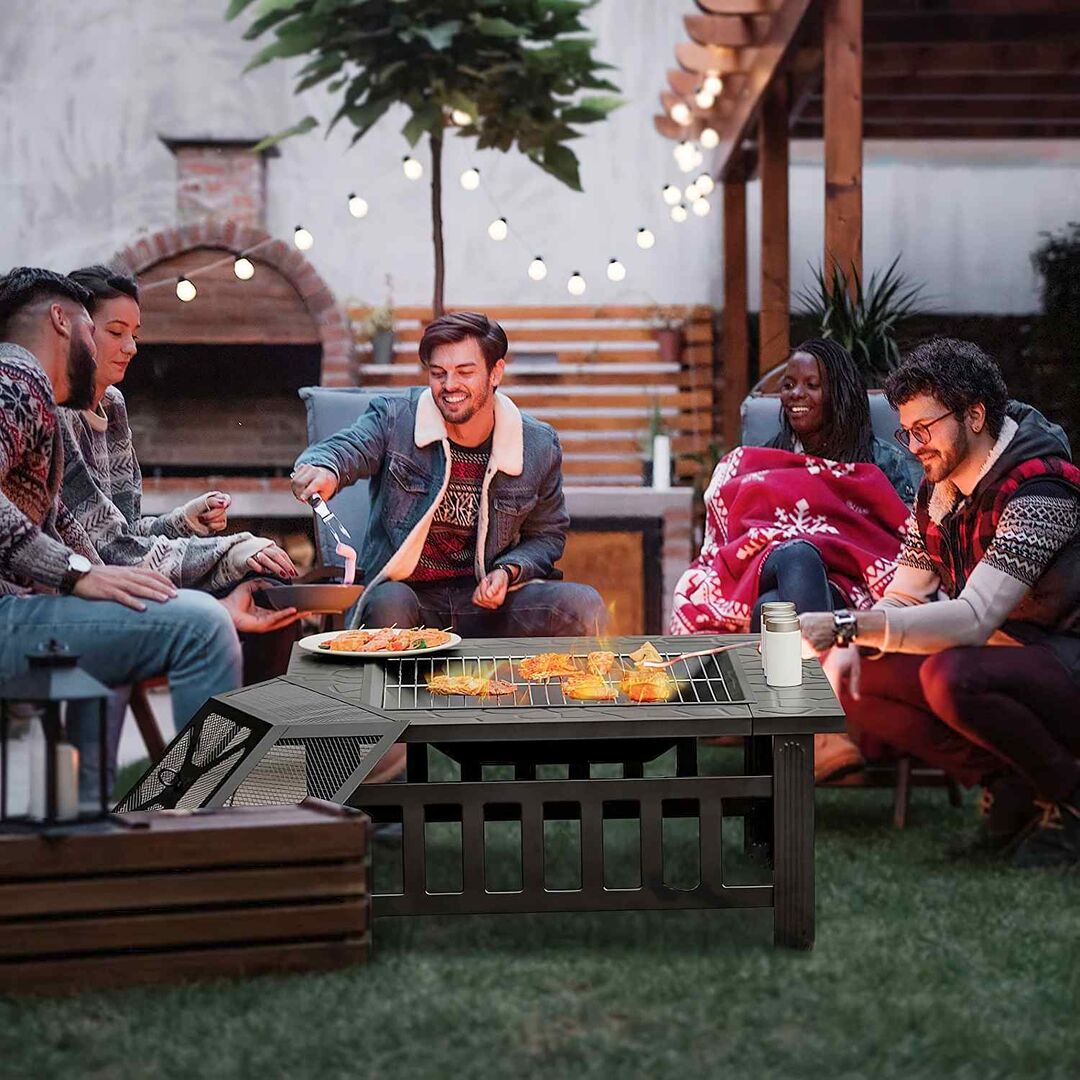 Gruppe von Freunden sitzt abends im Garten um eine Feuerstelle, grillt Essen und lacht gemeinsam; stimmungsvolle Beleuchtung mit Lichterkette im Hintergrund.