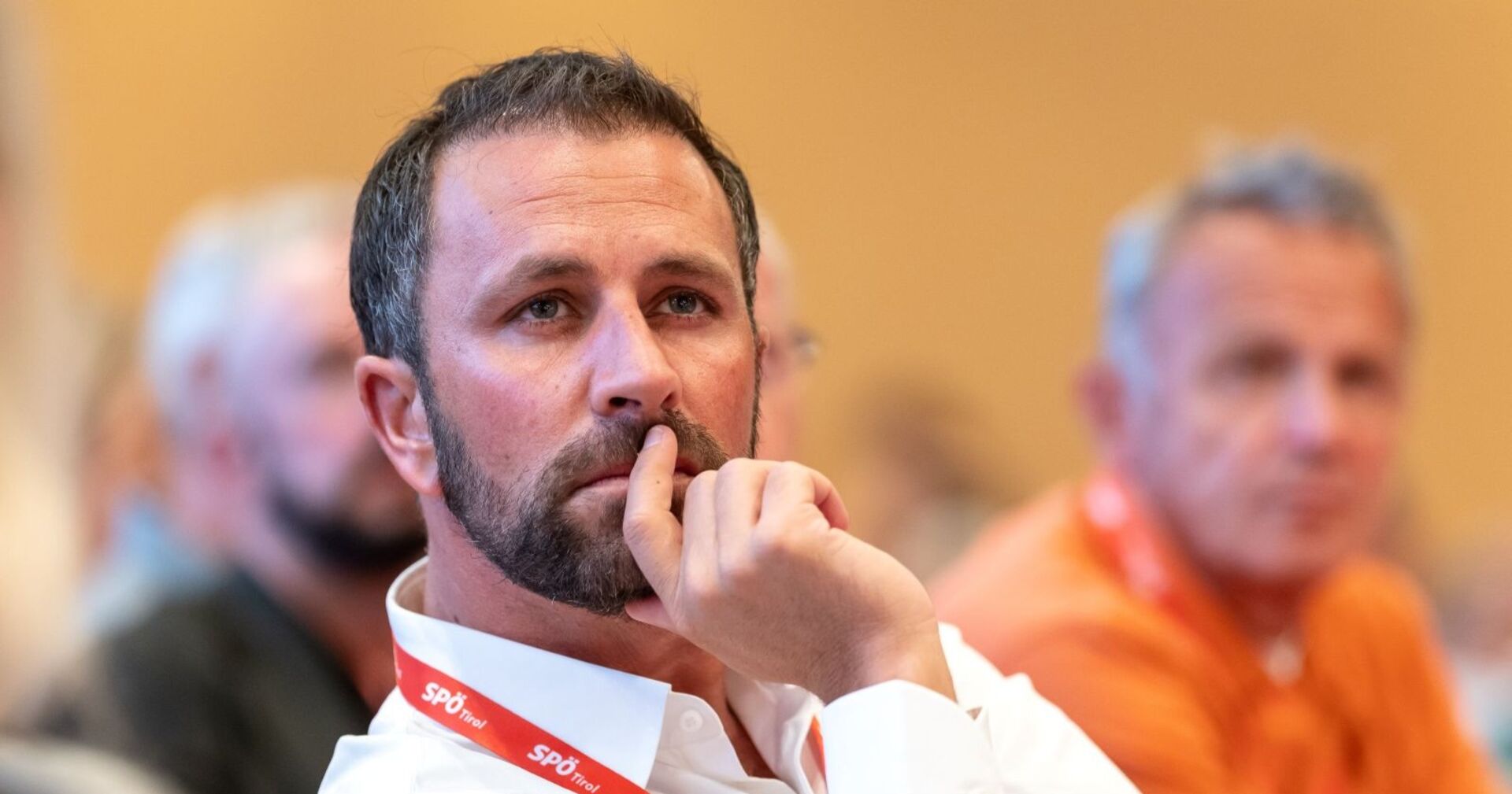 Georg Dornauer mit rotem SPÖ-Tirol-Lanyard sitzt nachdenklich bei einer Sitzung, Hand am Gesicht, unscharfe Teilnehmer im Hintergrund.