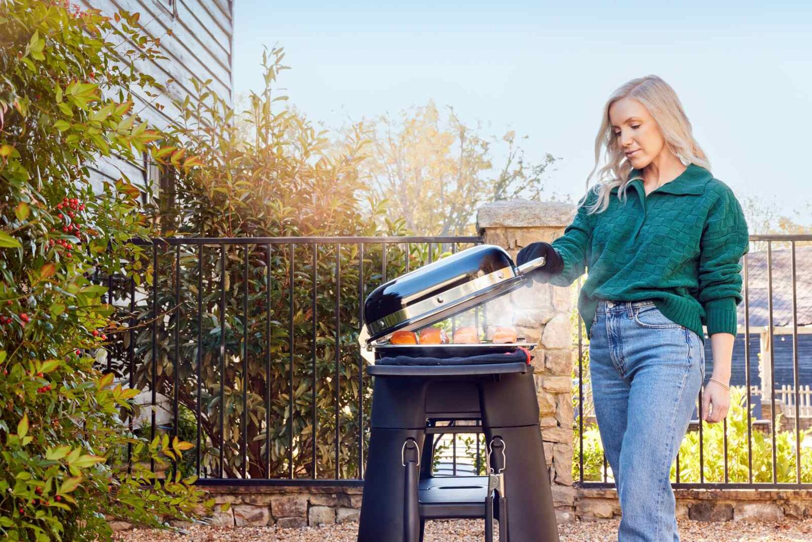 Frau grillt im Garten an einem modernen Gasgrill, trägt grünen Pullover und Jeans; sonniger Herbsttag mit Pflanzen, Steinmauer und entspannter Atmosphäre.