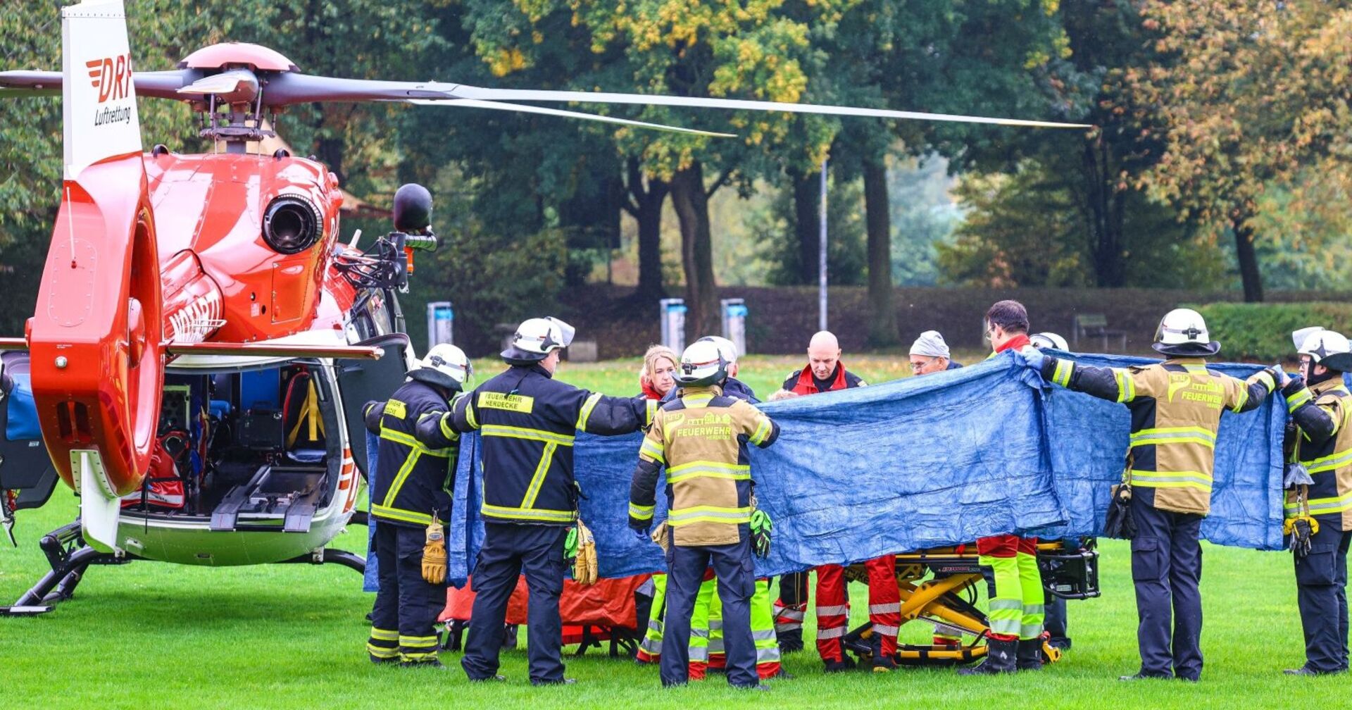 Feuerwehrleute und Sanitäter in Herdecke schirmen eine verletzte Person beim Transport zu einem roten Rettungshubschrauber ab.