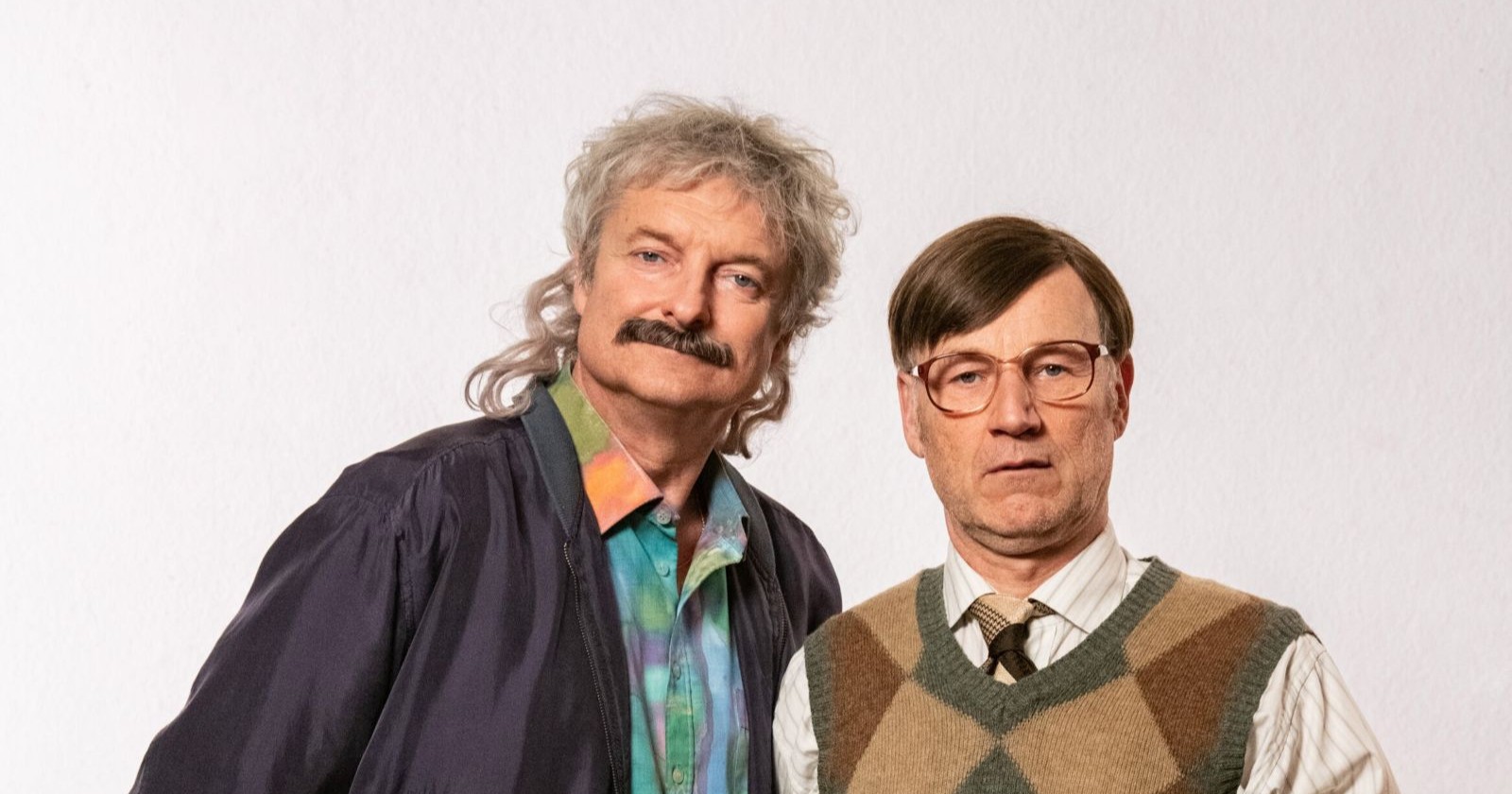 Zwei Männer in typischer 90er-Jahre-Aufmachung stehen nebeneinander vor hellem Hintergrund – das legendäre Comedy-Duo „Weber &amp; Breitfuß“ aus der ORF-Kultserie.