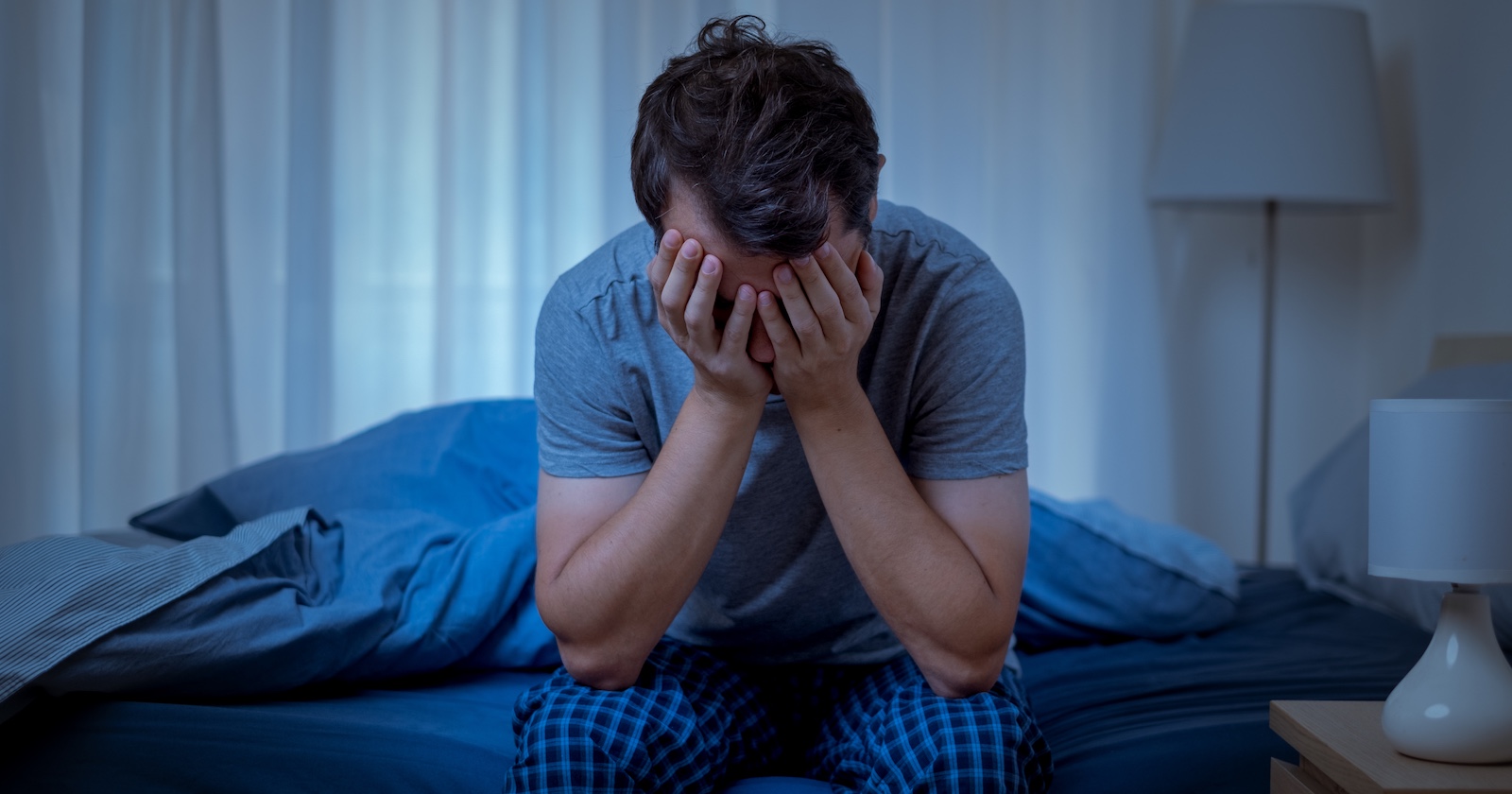 verzweifelter Mann sitzt im Pyjama am Bett und hält sich beide Hände vor das Gesicht