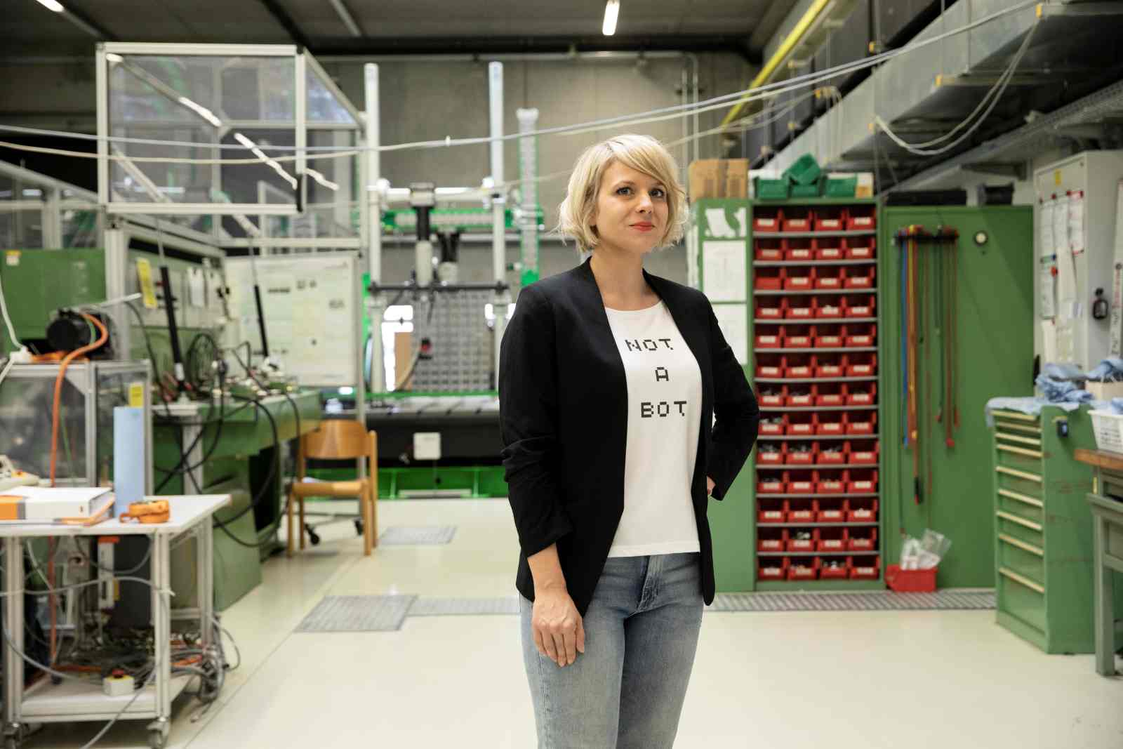 Eine Frau mit blondem, kurzem Haar steht in einer Werkhalle oder einem Labor. Sie trägt ein weißes T-Shirt mit der Aufschrift „NOT A BOT“, dazu eine schwarze Jacke und Jeans. Im Hintergrund sind technische Geräte, Werkbänke und Werkzeuge zu sehen.