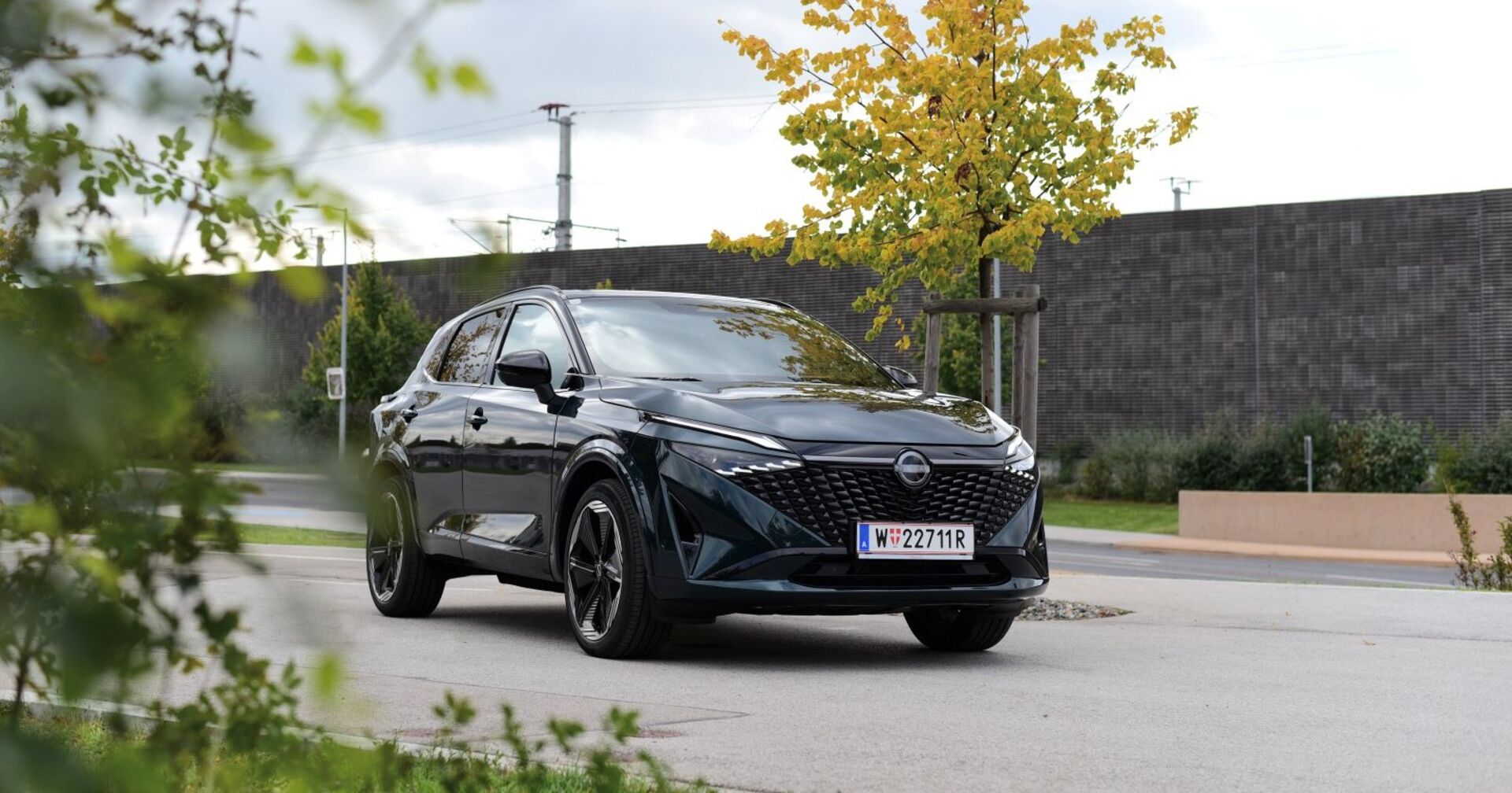 Ein schwarzer   Nissan Qashqai steht vor auf einer Straße. Im Hintergrund ist eine Schallschutzmauer aus Holz und ein herbstlich gefärbter Laubbaum zu sehen.