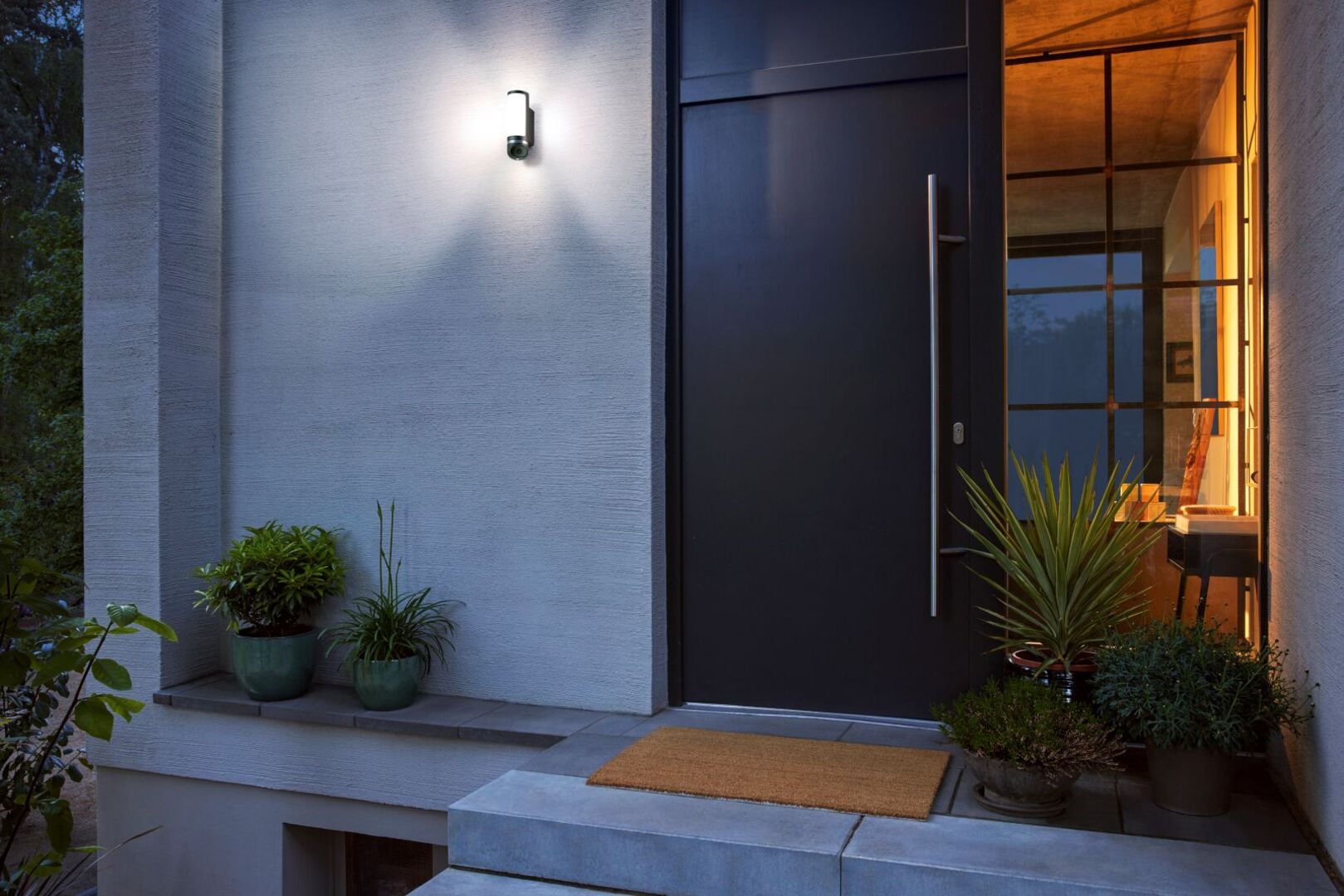 Moderne Haus­eingangsszene mit beleuchteter Außenlampe und integrierter Sicherheitskamera neben einer schwarzen Haustür, umgeben von grünen Pflanzen am Eingang.