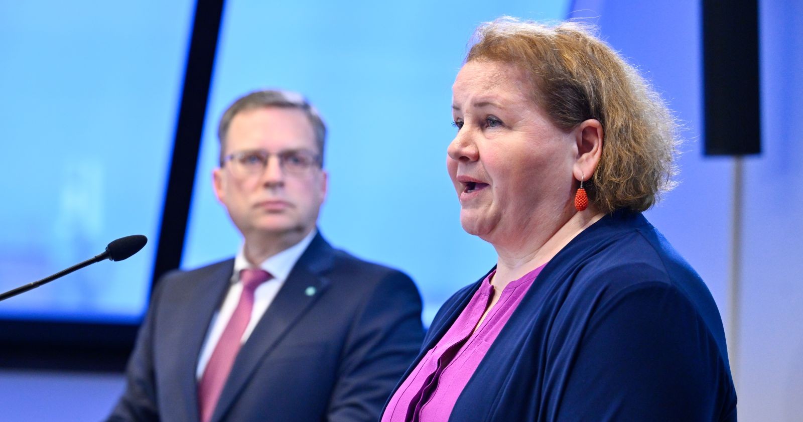 Sozialministerin steht bei einer Pressekonferenz am Rednerpult, im Hintergrund blickt Wöginger zu ihr.