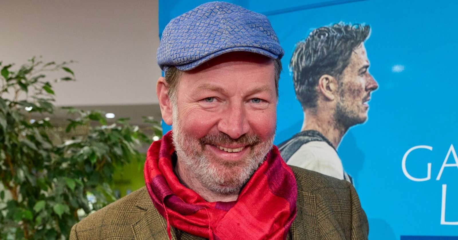 Robert Reinagl lächelt bei einer Veranstaltung, trägt eine blaue Kappe und einen roten Schal, im Hintergrund ist ein Plakat zu sehen.