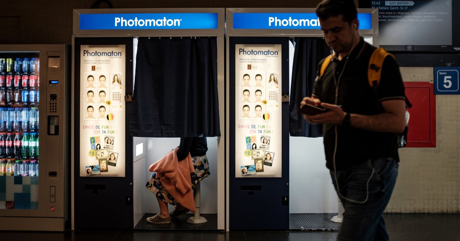 Fotobox im Bahnhofsgebäude, teilweise geschlossener Vorhang, eine Person sitzt drinnen, daneben ein Passant mit Rucksack – Symbolbild für den Vorfall am Rostocker Hauptbahnhof.