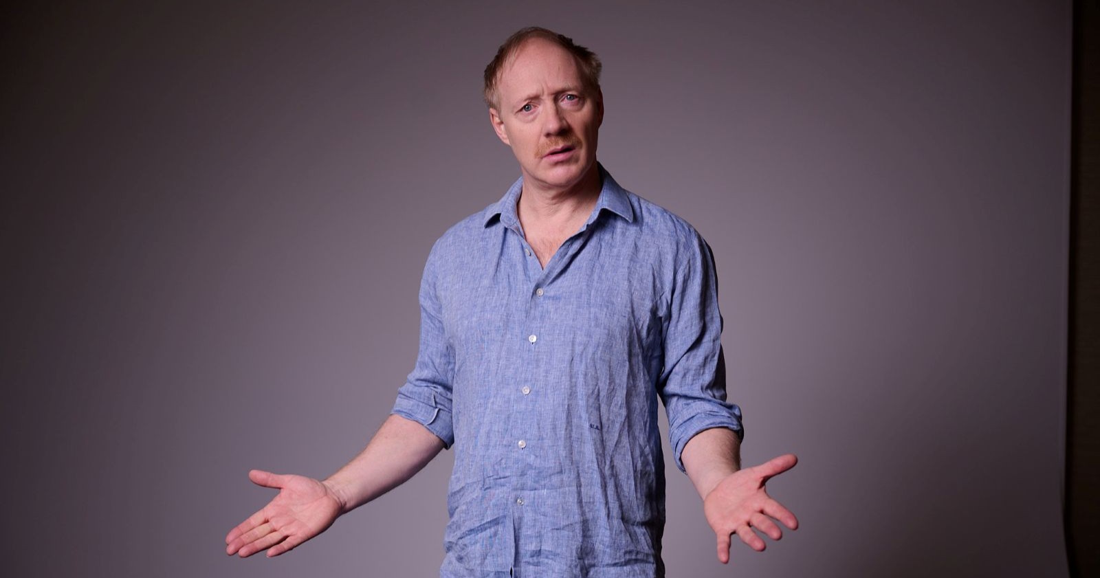 Schauspieler Simon Schwarz steht in blauem Stoffhemd vor einer Schwarzen Leinwand mit einem fragenden Blick