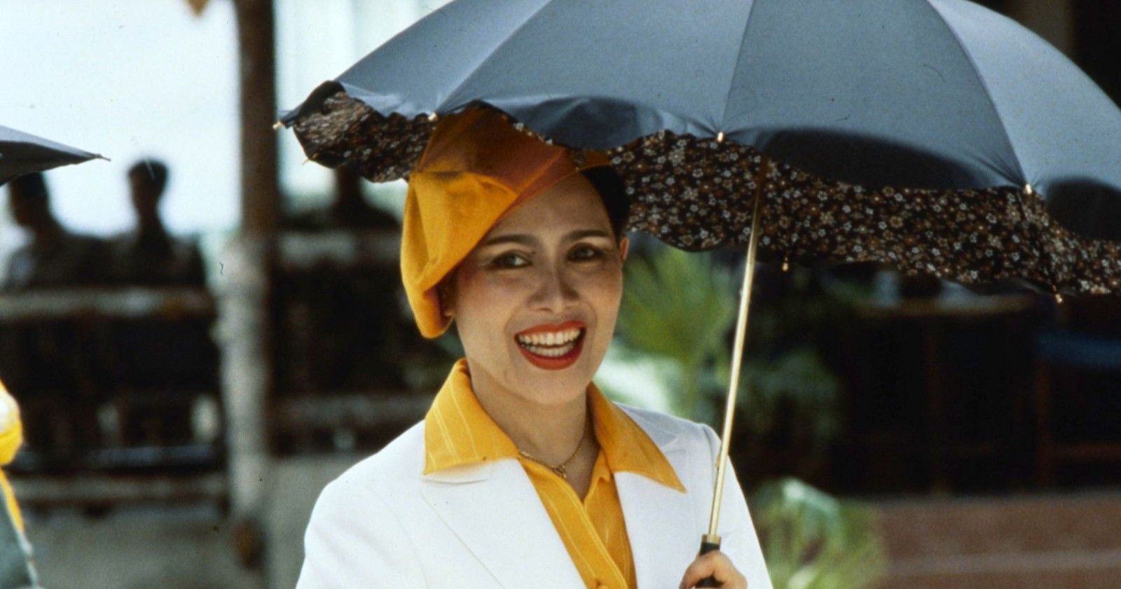 Queen Sirikit trägt ein gelbes Barett und hält einen Schirm; sie lächelt in die Kamera