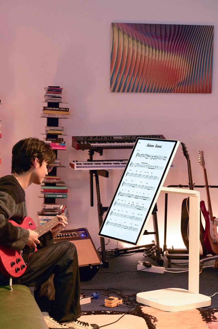 Person spielt E-Gitarre im Musikzimmer, vor ihr steht ein vertikal ausgerichteter Smart Monitor mit digital angezeigten Noten; im Hintergrund Musikinstrumente und Bücher.