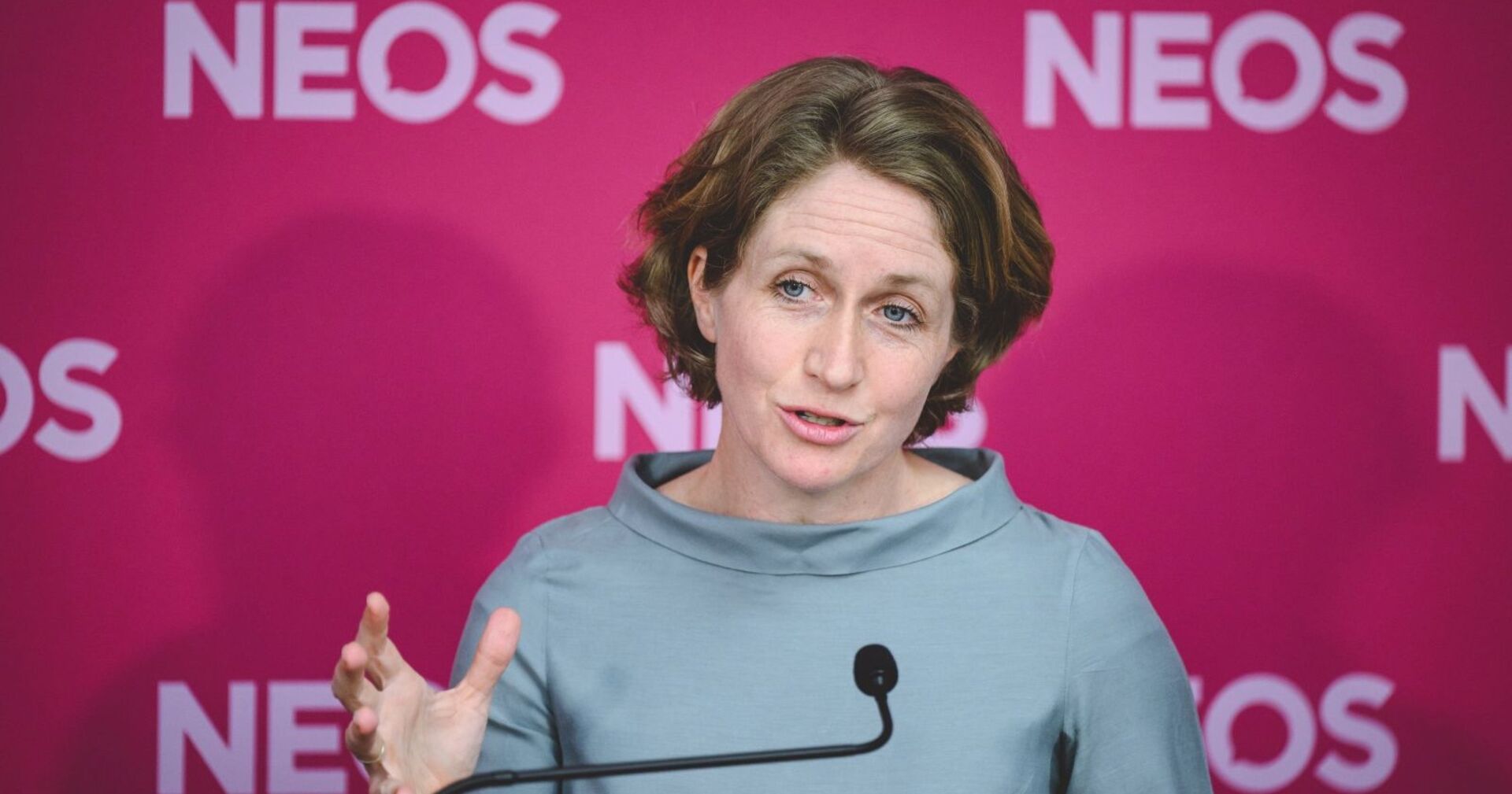 Stephanie Krisper steht vor einem pinken NEOS-Hintergrund und spricht bei einer Pressekonferenz, während sie eine Handgeste macht. Jetzt hat sie ihr Mandat zurückgelegt.