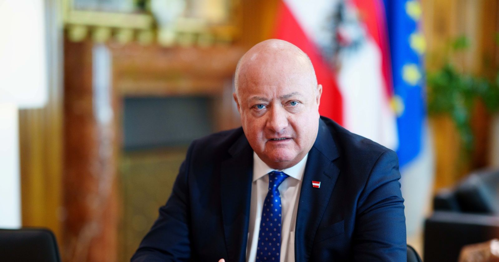 Bundeskanzler Stocker vor seiner Rücken-Operation in dunklem Anzug und blauer Krawatte sitzt im Büro vor Österreich- und EU-Flagge.