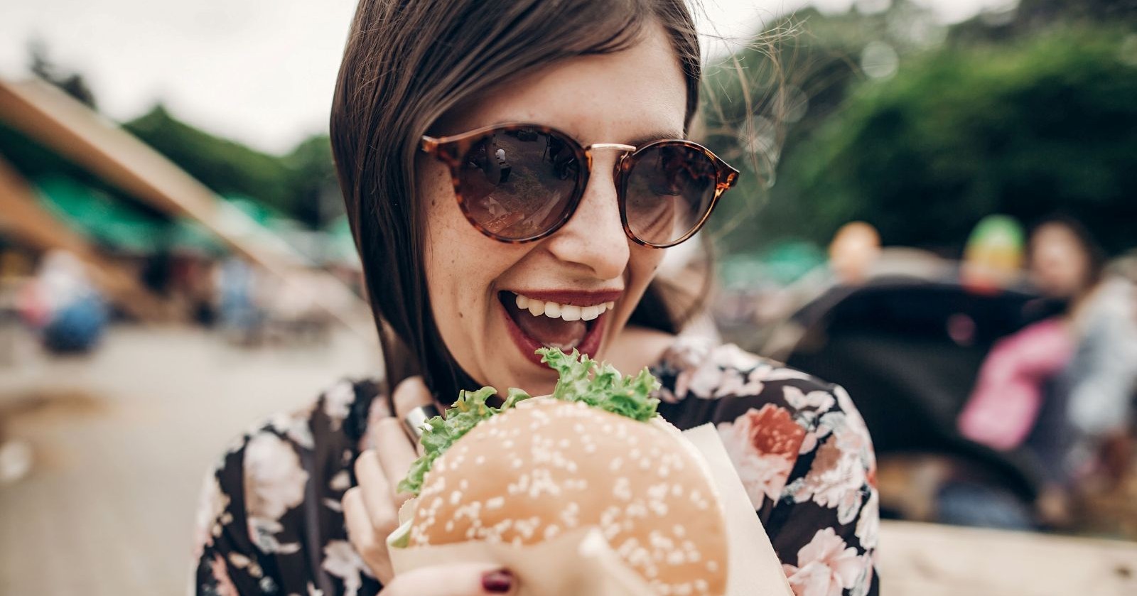 Frau mit Sonnenbrille hält lächelnd einen Burger auf einem Street-Food-Festival in der Hand.