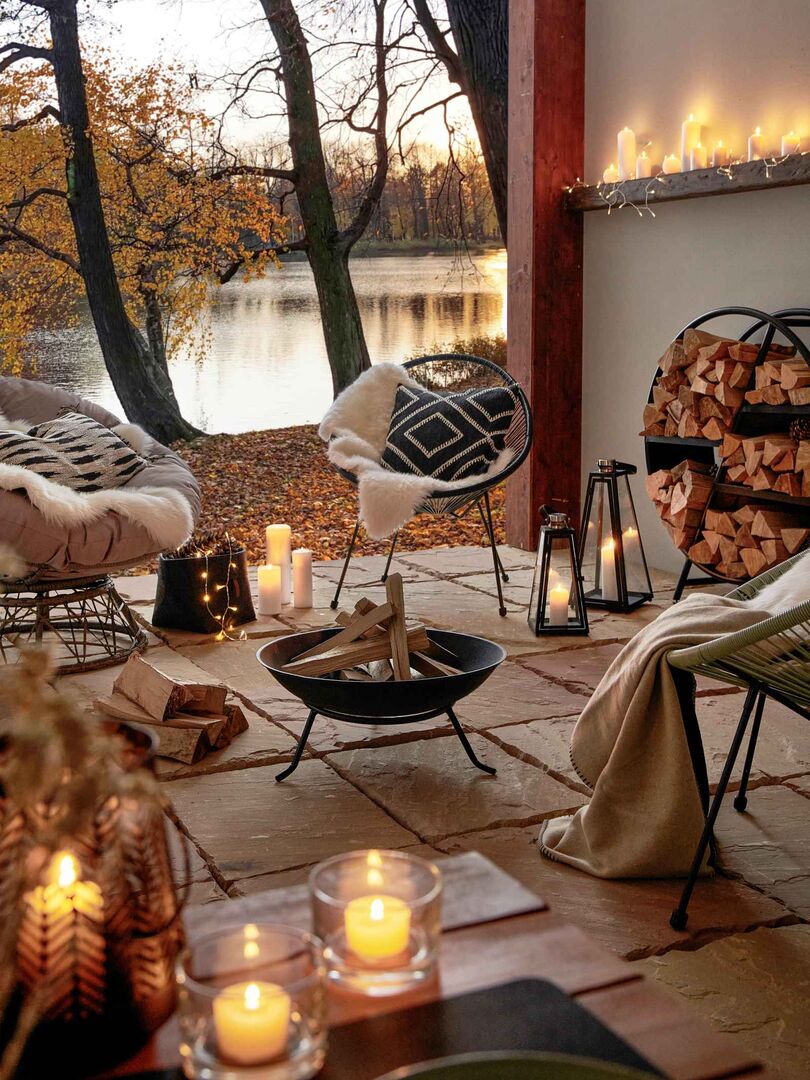 Gemütlich dekorierte Terrasse im Herbst mit Kerzen, Feuerstelle, Fellen und Decken, Blick auf einen See mit buntem Laub – stimmungsvolle Outdoor-Atmosphäre.
