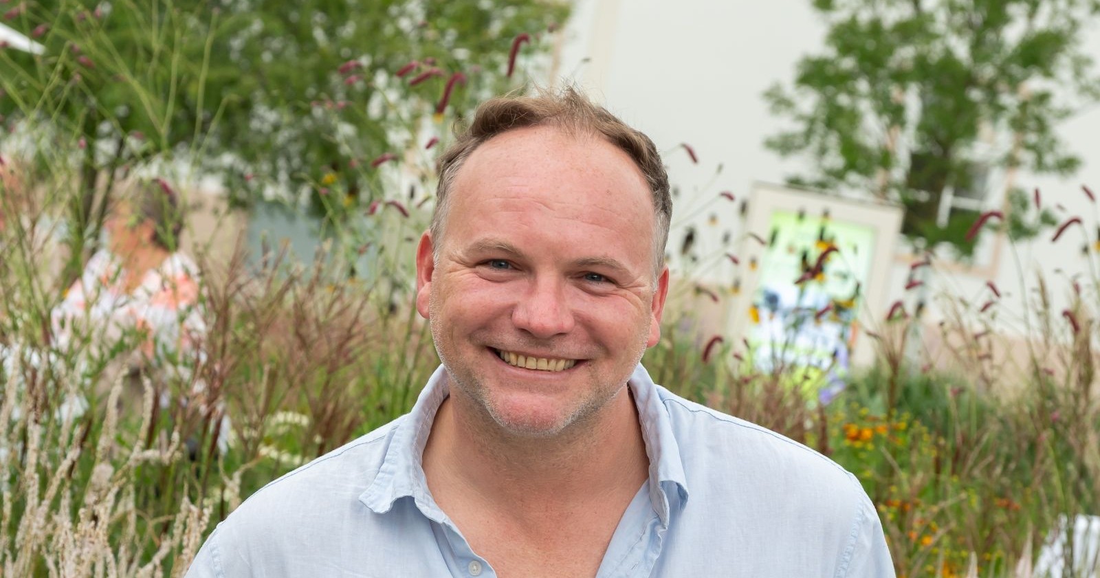 Kabarettist Thomas Stipsits lächelt in die Kamera, im Hintergrund blühende Pflanzen und Sommerstimmung.