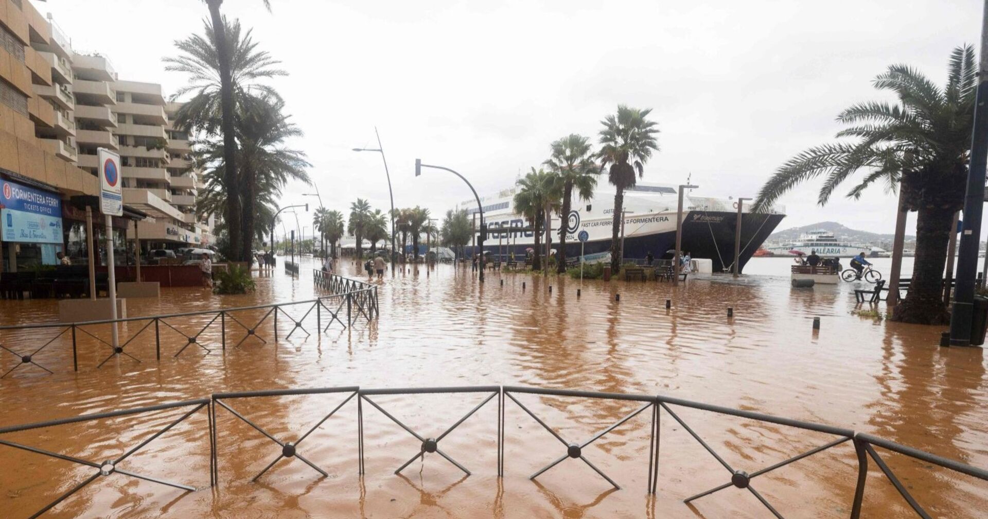 Überflutete Straßen im Hafen von Ibiza Stadt mit stehenden Wassermassen, Palmen und festliegenden Fähren im Hintergrund.