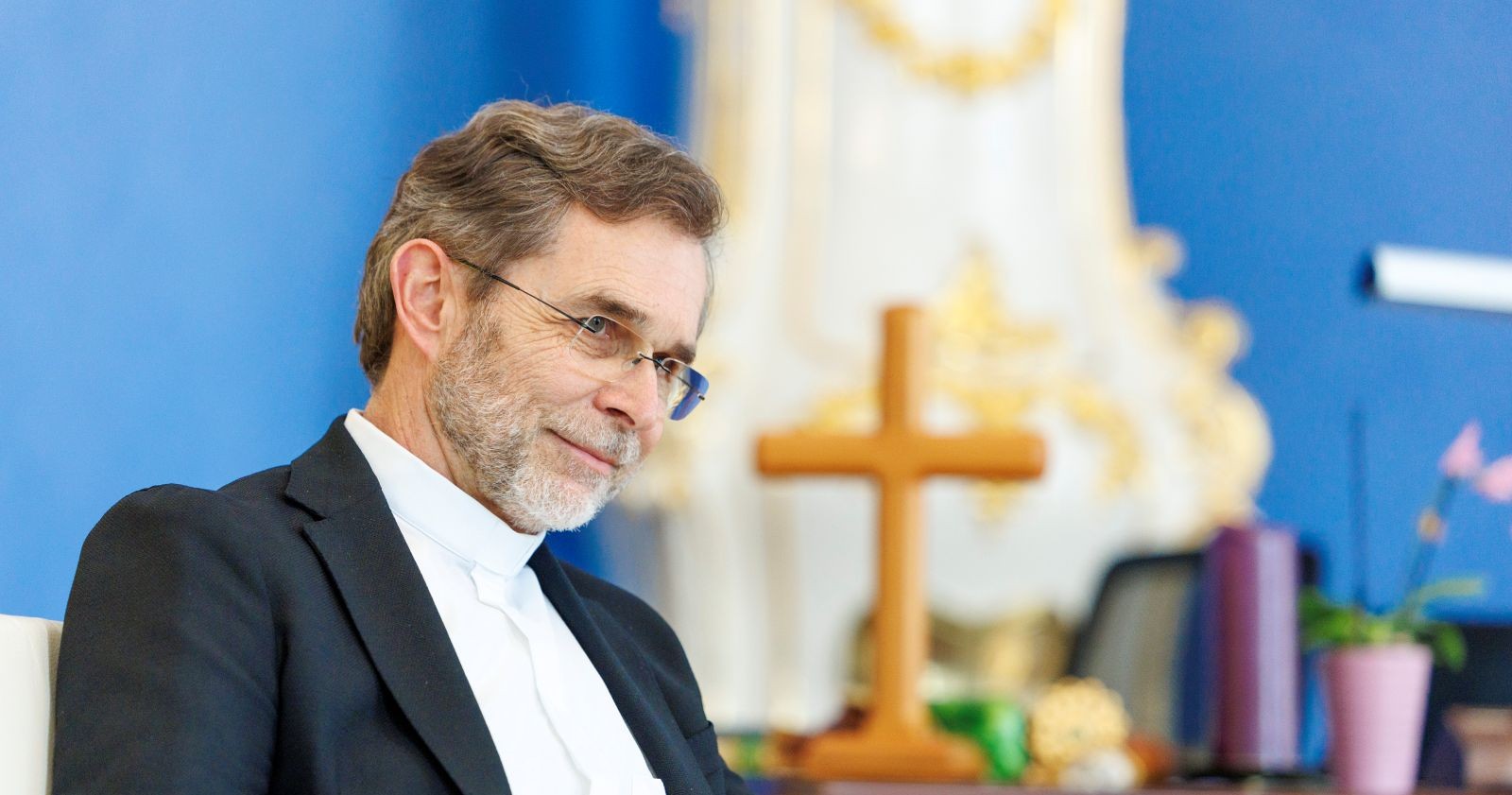Josef Grünwidl freundlich lächelnd mit Brille sitzt vor blauem Hintergrund, hinter ihm steht ein Holzkreuz und eine Blumenvase.