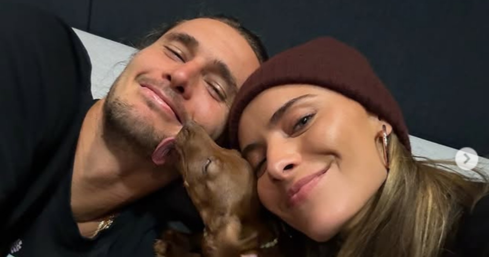 Zverev und Thomalla liegen lächelnd nebeneinander, während ihr kleiner brauner Dackelwelpe ihnen liebevoll über das Gesicht leckt.